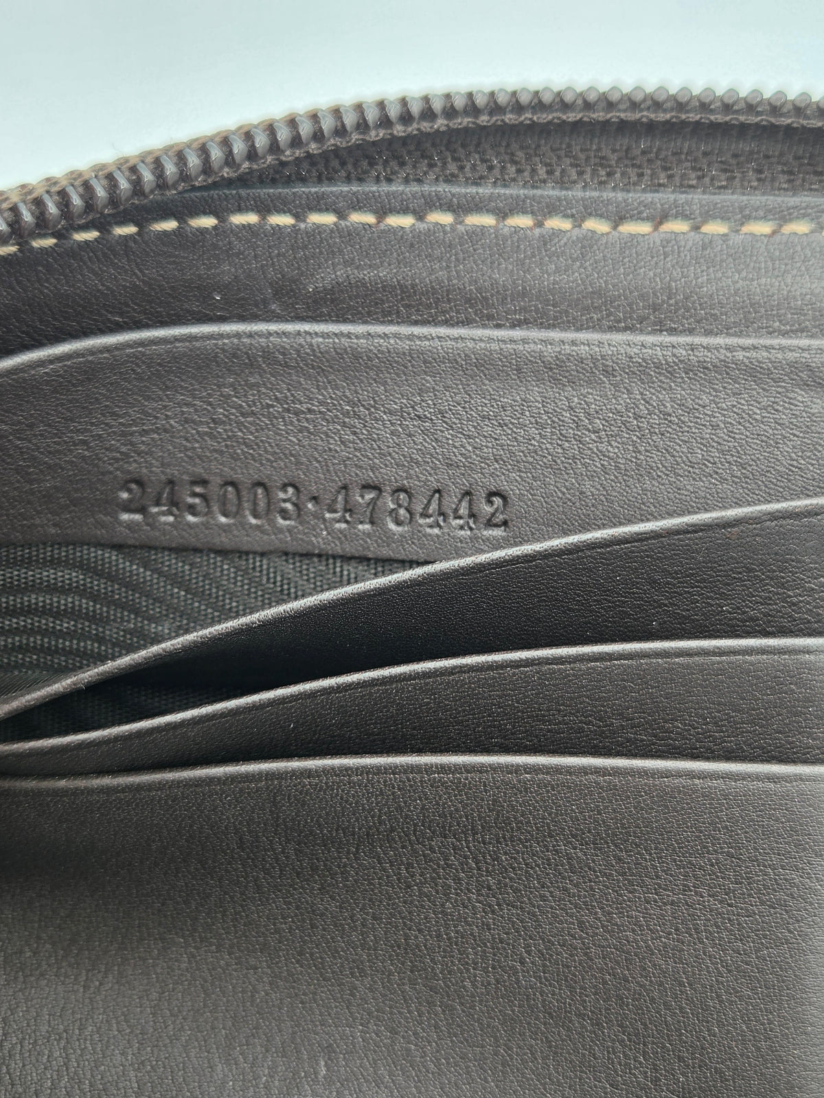 Close-up of the interior of a dark gray leather wallet, showing card slots, a zipper edge with tan stitching, and embossed serial numbers '245003-478442'.