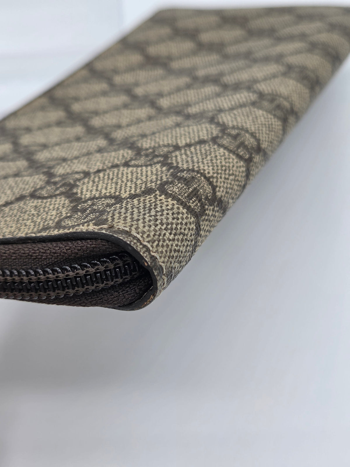 A close-up of a long wallet or clutch in brown and beige GG Supreme canvas with a dark brown zipper along the edge, shown on a light gray background.