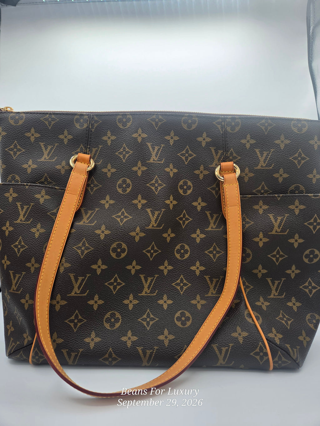 A Louis Vuitton monogram canvas tote bag with tan leather handles and gold-tone hardware, photographed against a light gray background.
