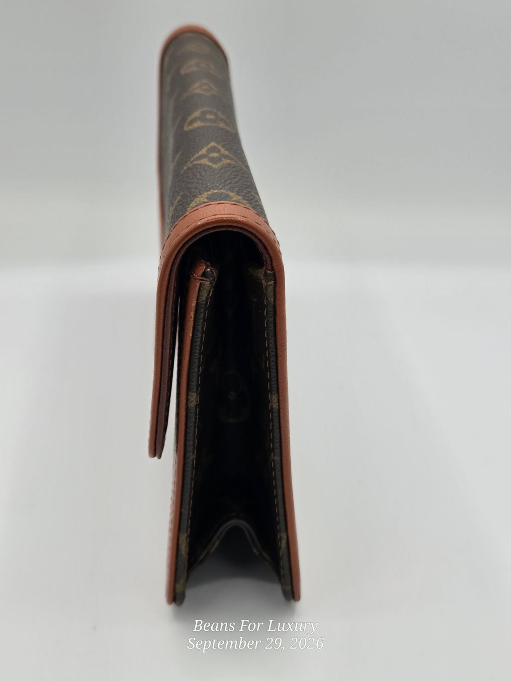 A close-up side view of a brown monogram wallet or card holder with tan leather trim, photographed against a light gray background.
