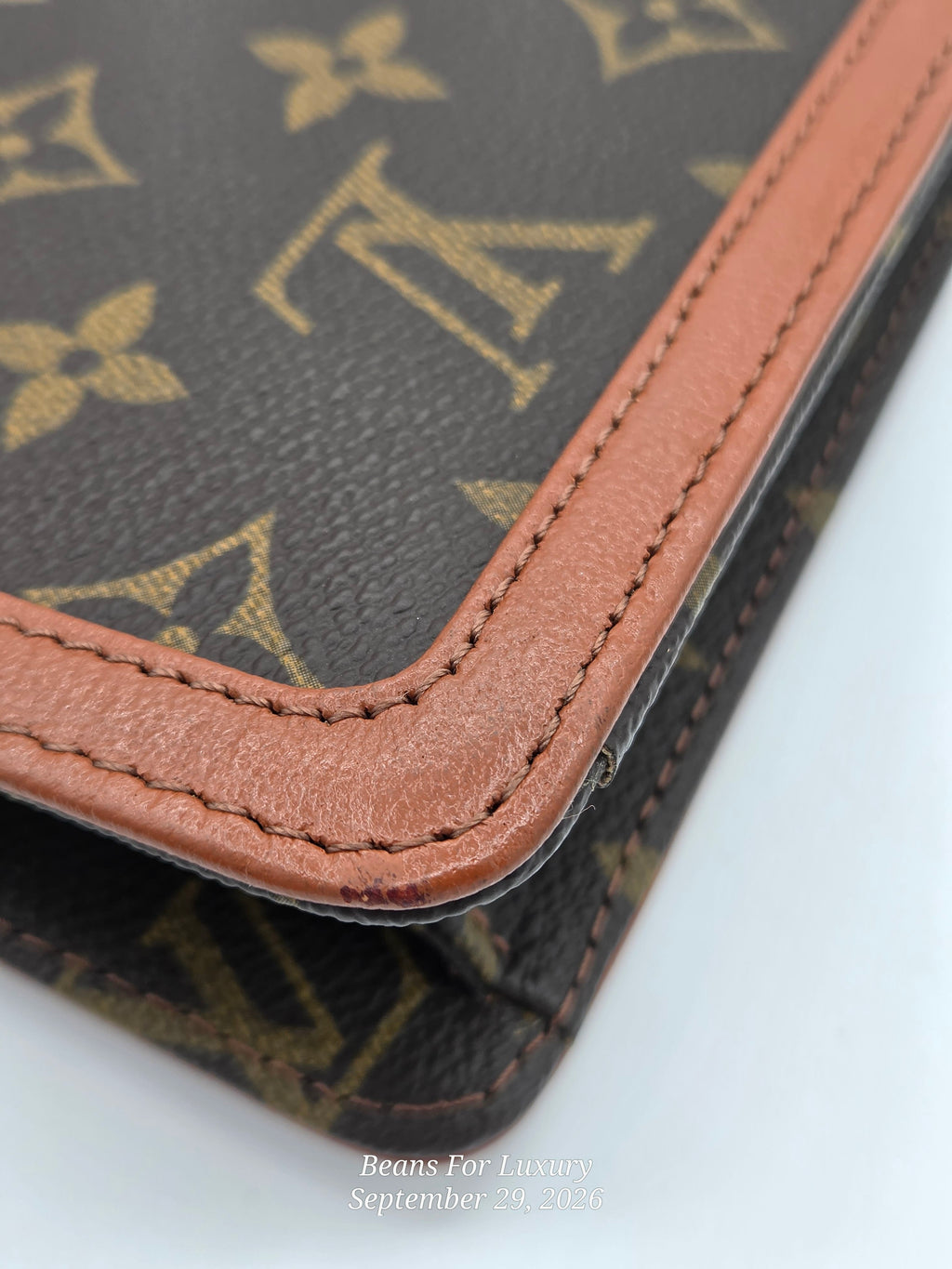 Close-up of a Louis Vuitton monogram wallet or notebook corner showing dark brown coated canvas with tan monogram print and contrasting tan leather trim, photographed on a white background.