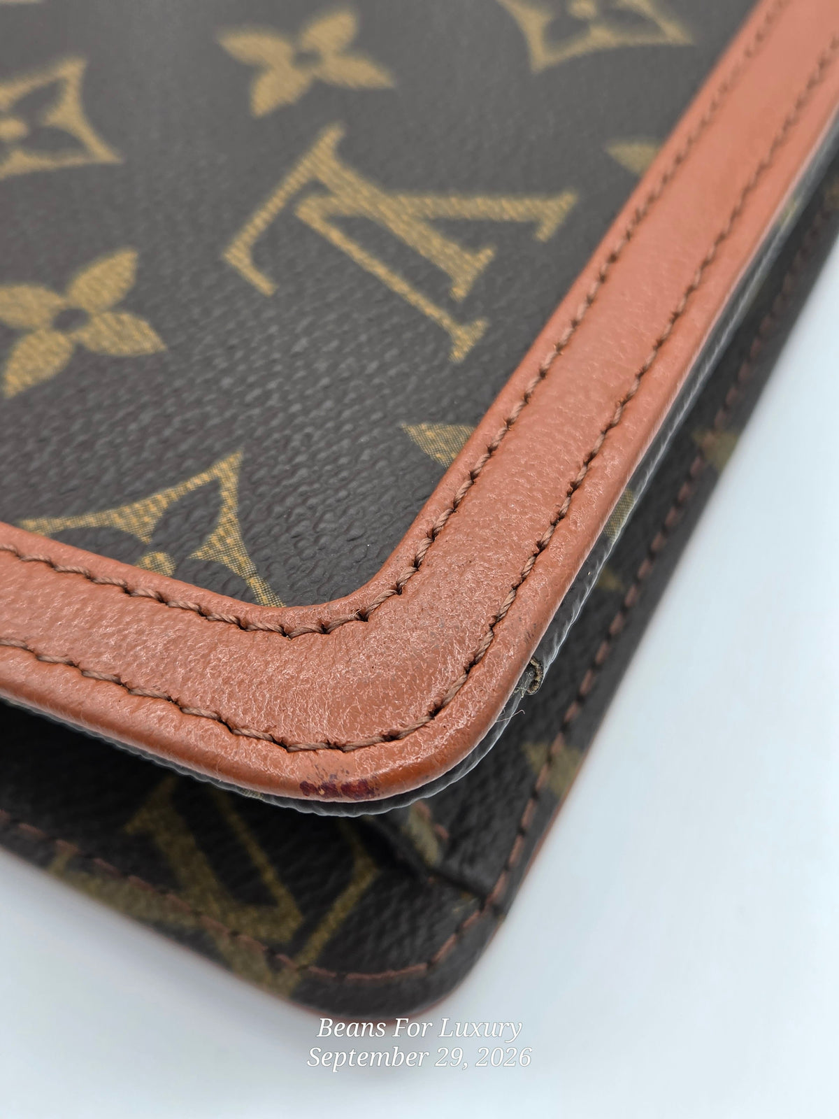 Close-up of a Louis Vuitton monogram wallet or notebook corner showing dark brown coated canvas with tan monogram print and contrasting tan leather trim, photographed on a white background.