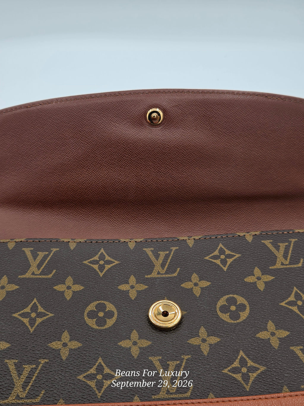 Close-up of a Louis Vuitton monogram clutch or envelope bag showing the brown coated canvas exterior with gold-tone hardware and a brown leather interior flap. Visible LV monogram pattern and a small watermark reading Beans For Luxury September 29, 2026.