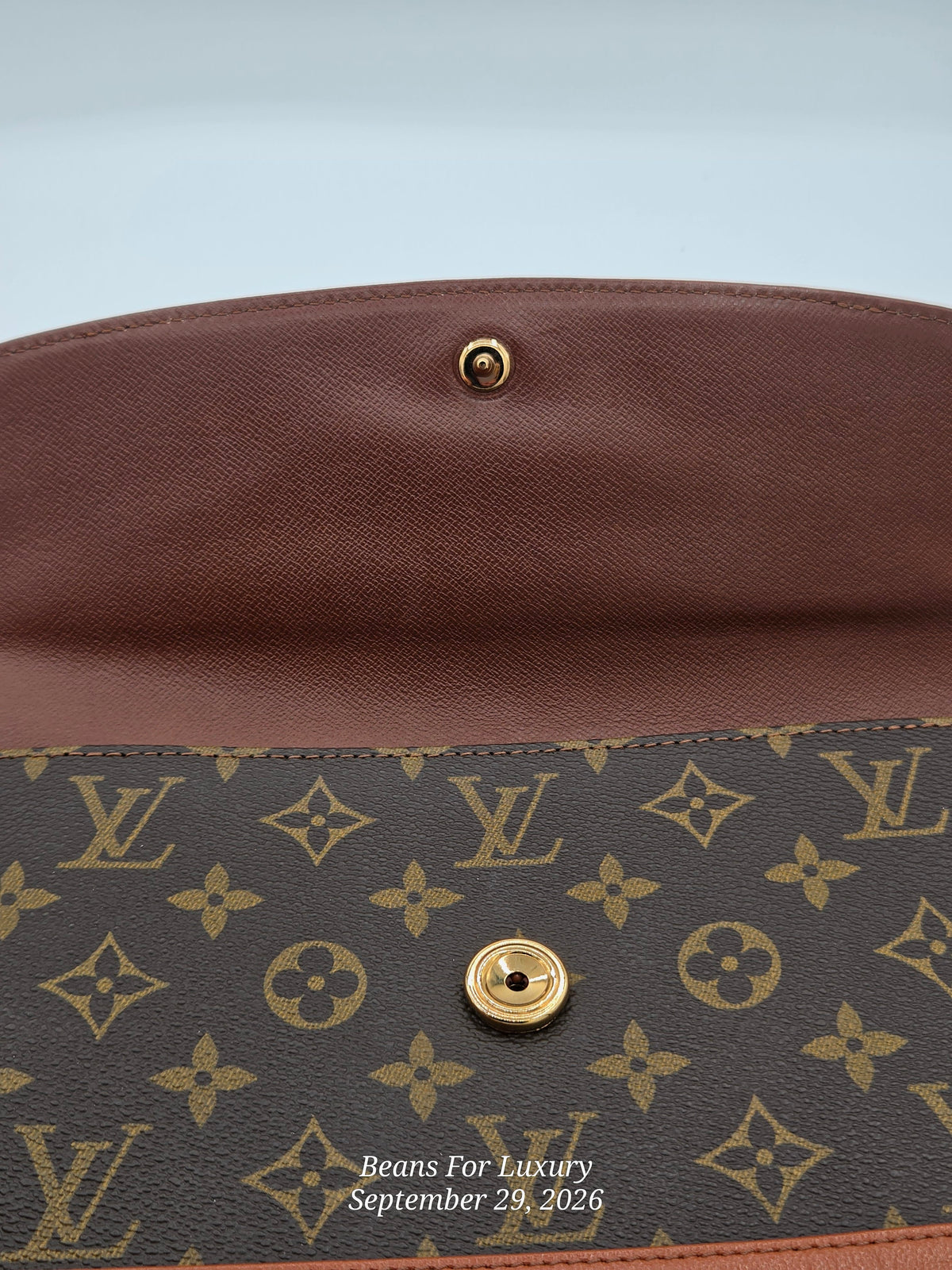 Close-up of a Louis Vuitton monogram clutch or envelope bag showing the brown coated canvas exterior with gold-tone hardware and a brown leather interior flap. Visible LV monogram pattern and a small watermark reading Beans For Luxury September 29, 2026.