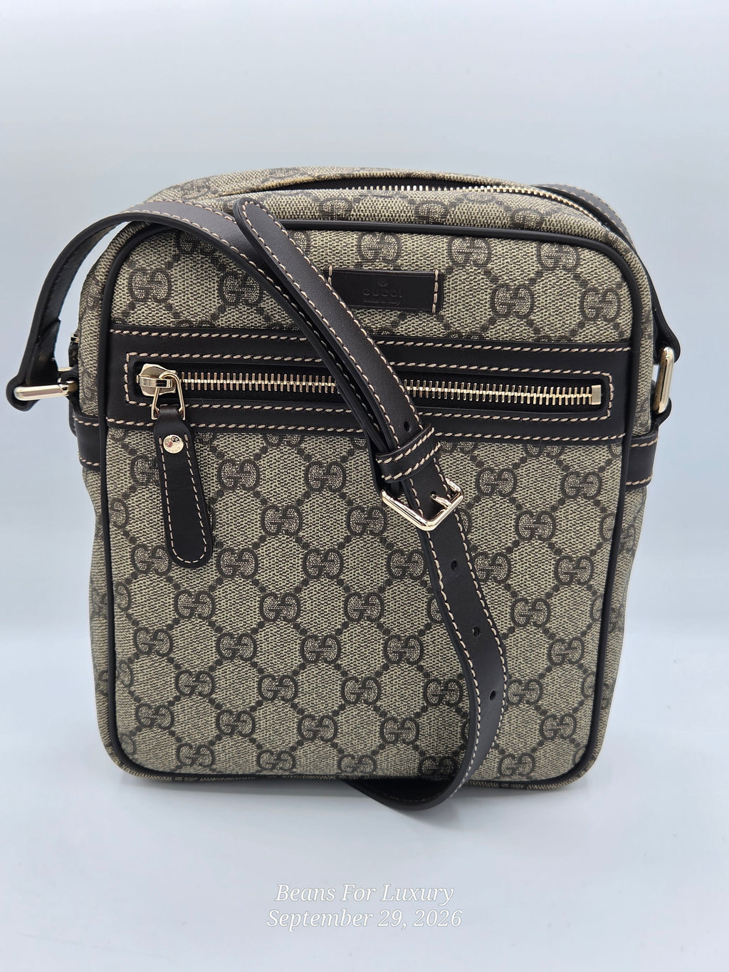 A Gucci crossbody bag in the classic GG Supreme canvas, featuring a beige and brown monogram pattern with dark brown leather trim and gold-tone hardware. The bag is shown against a light gray background with a Gucci label visible on the front.