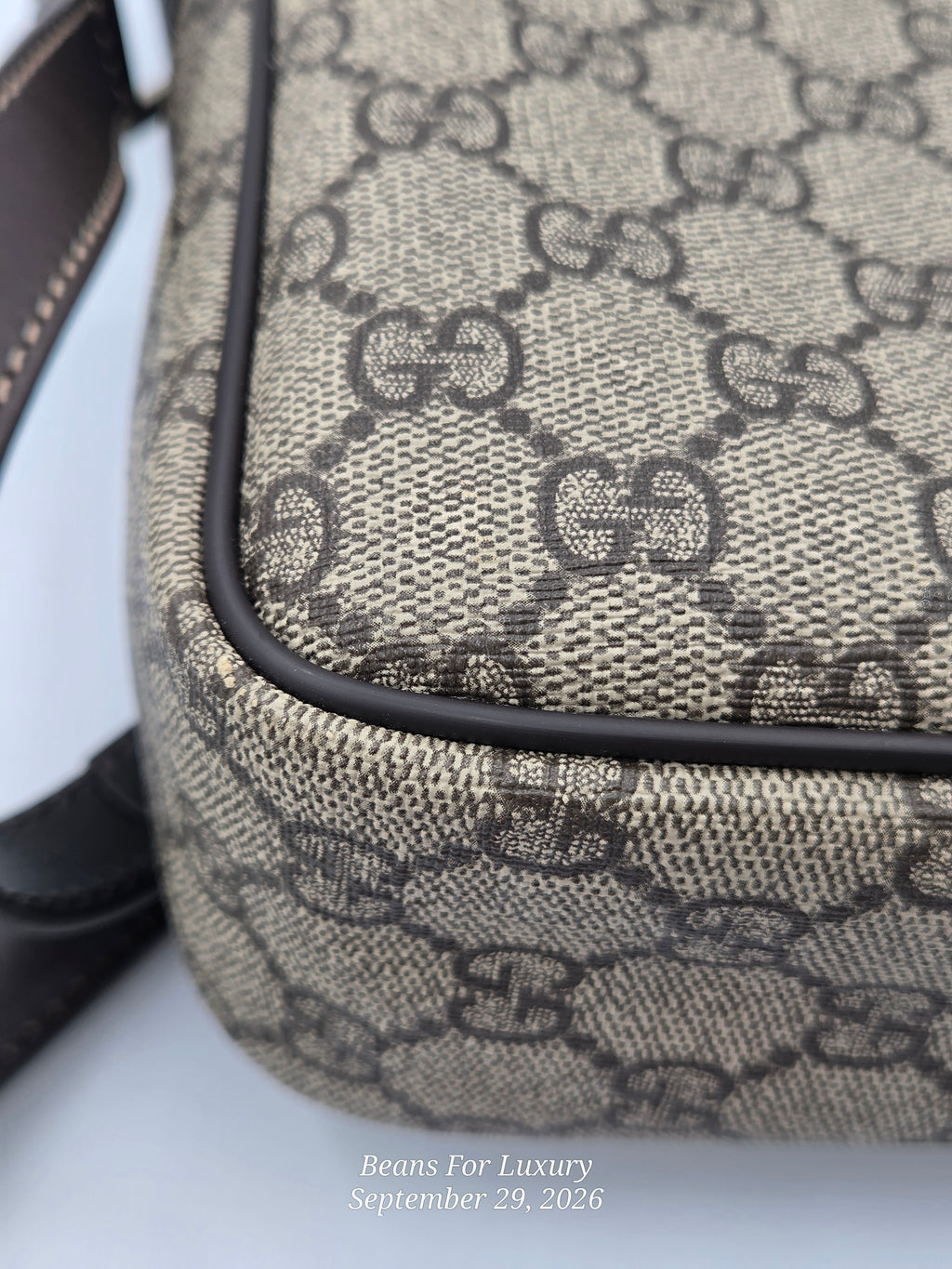 Close-up of a beige and brown monogram canvas bag corner with black piping and a dark strap edge, on a light background. Small watermark text at the bottom reads Beans For Luxury September 29, 2026.