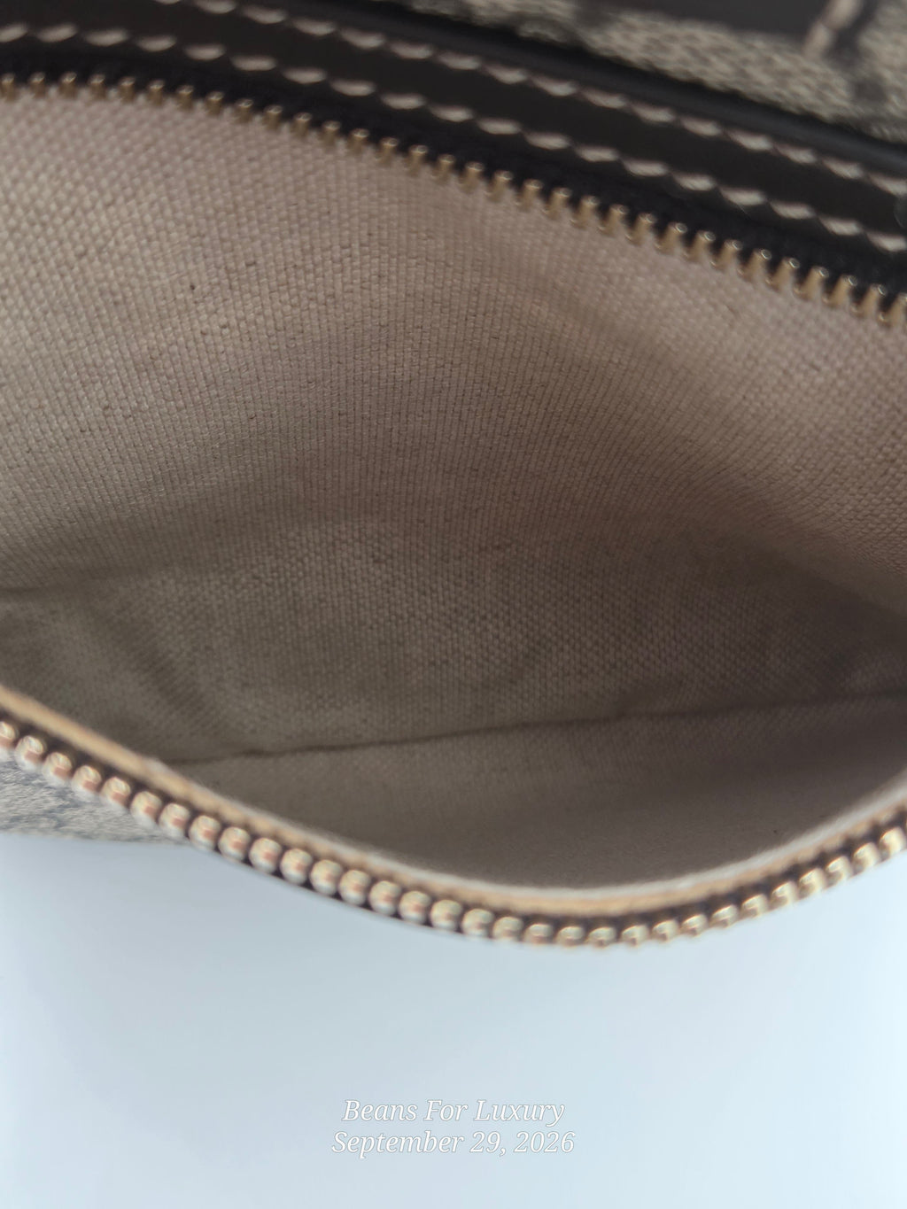 Close-up of the interior of a beige canvas zippered pouch or bag, showing the tan fabric lining, black stitched trim, and metallic zipper.