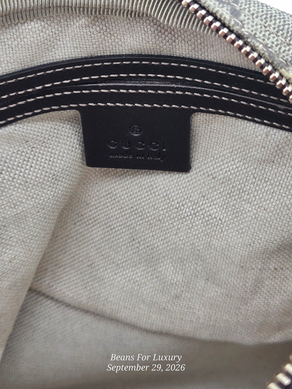 Close-up of the interior of a Gucci bag showing a black leather brand patch with white stitching, a light beige canvas body, and part of a metallic zipper.