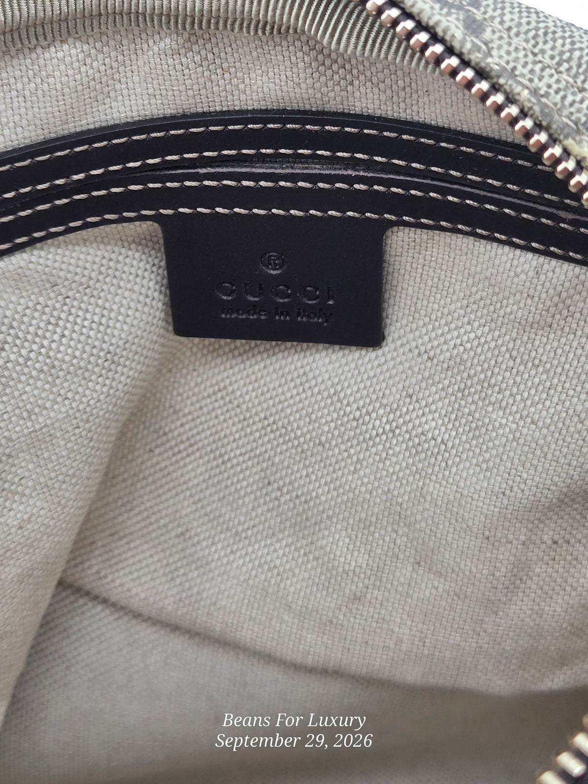 Close-up of the interior of a Gucci bag showing a black leather brand patch with white stitching, a light beige canvas body, and part of a metallic zipper.