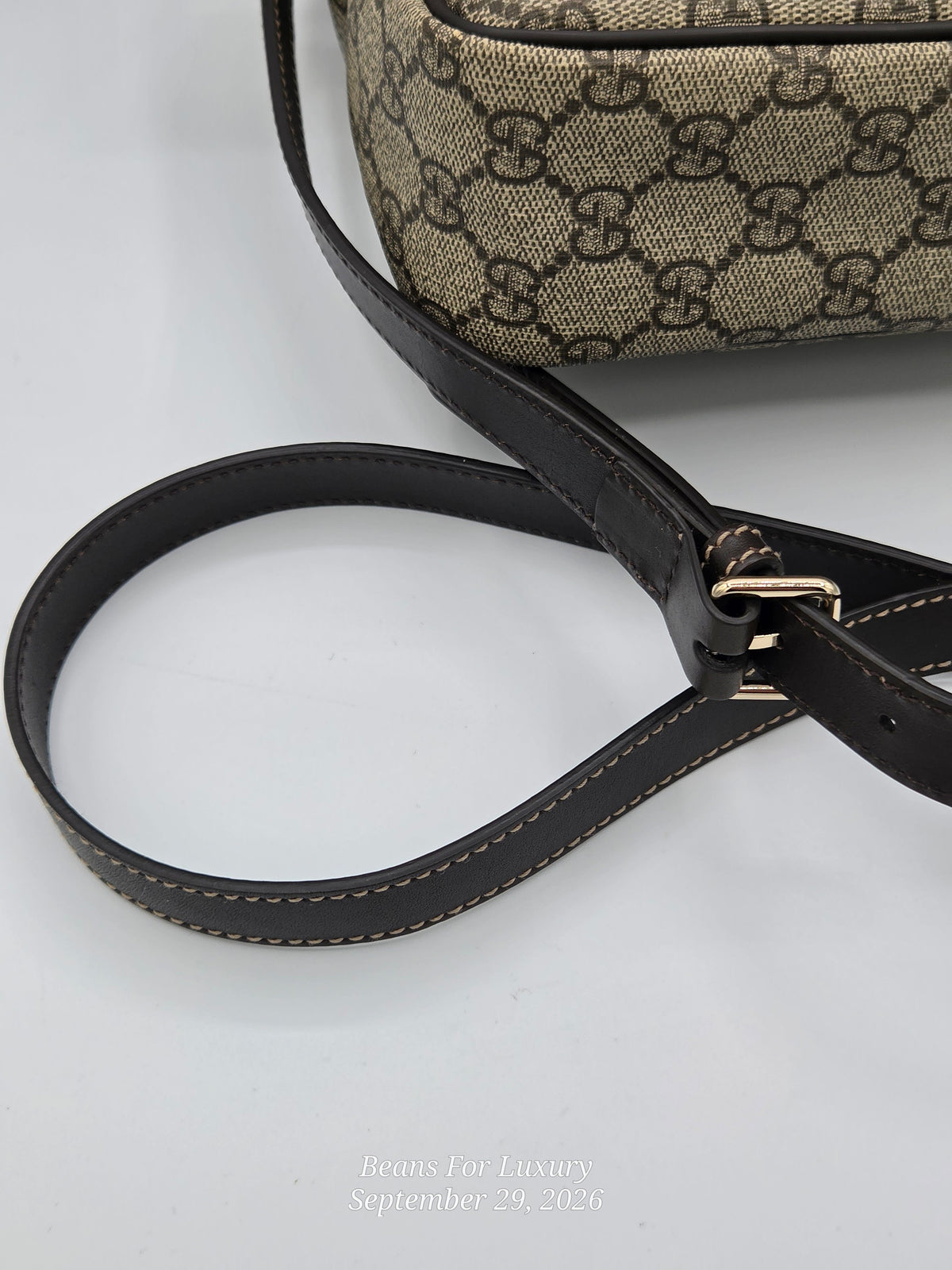 Close-up of a Gucci monogram handbag strap with black leather and gold-tone hardware, shown against a light gray background.