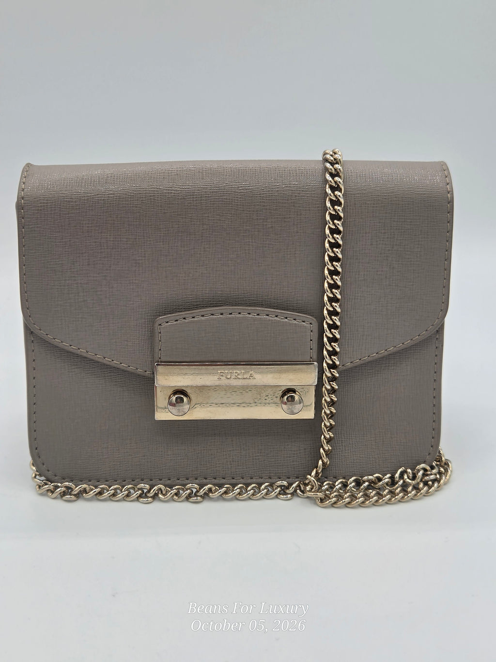 A taupe-brown textured leather crossbody bag with a gold-tone metal clasp and a gold-tone chain strap, shown on a light gray background.