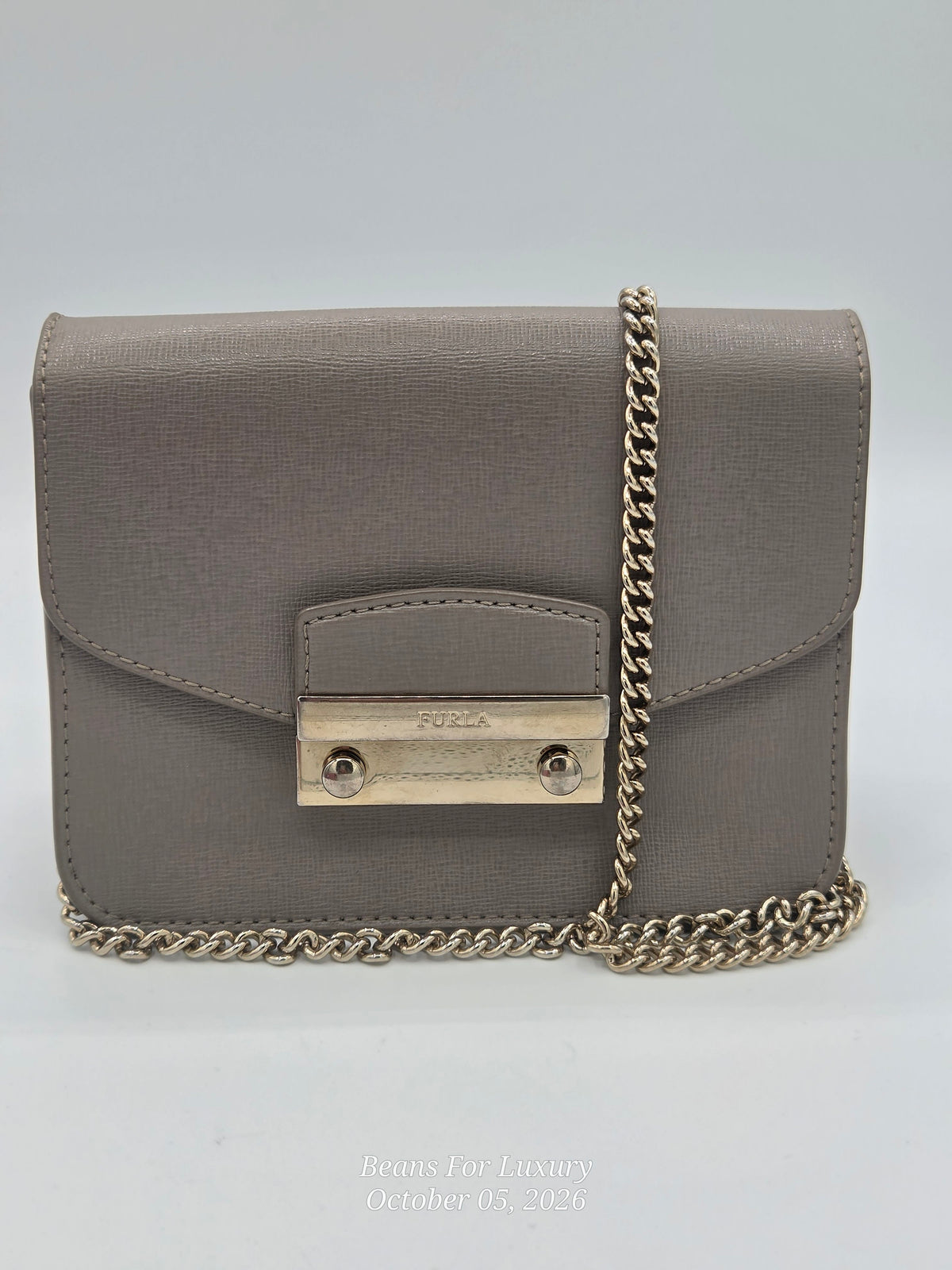 A taupe-brown textured leather crossbody bag with a gold-tone metal clasp and a gold-tone chain strap, shown on a light gray background.