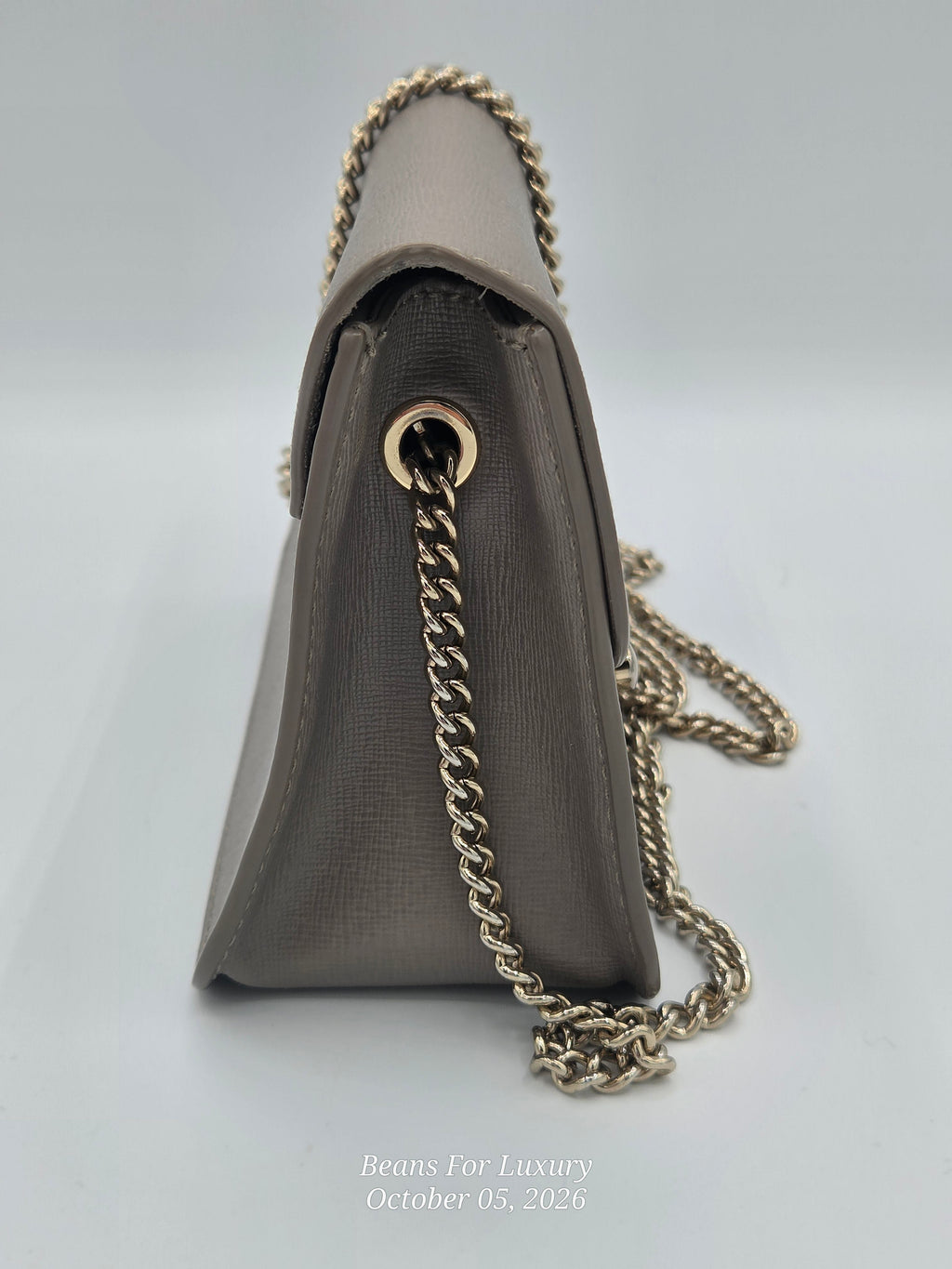 A taupe-brown textured leather shoulder bag shown from a side angle, featuring a gold-tone chain strap and a gold-tone grommet detail, set against a light gray background.