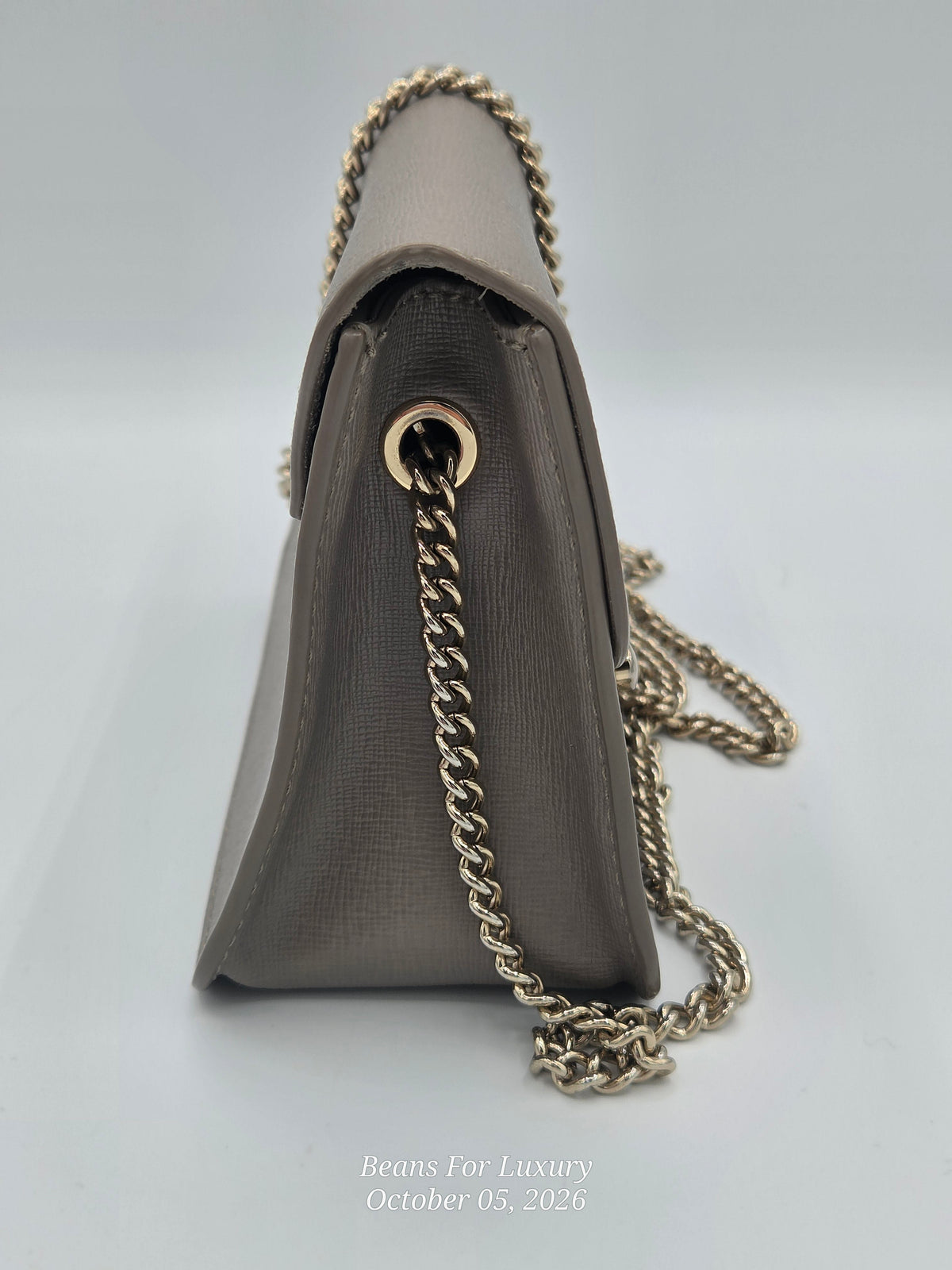 A taupe-brown textured leather shoulder bag shown from a side angle, featuring a gold-tone chain strap and a gold-tone grommet detail, set against a light gray background.