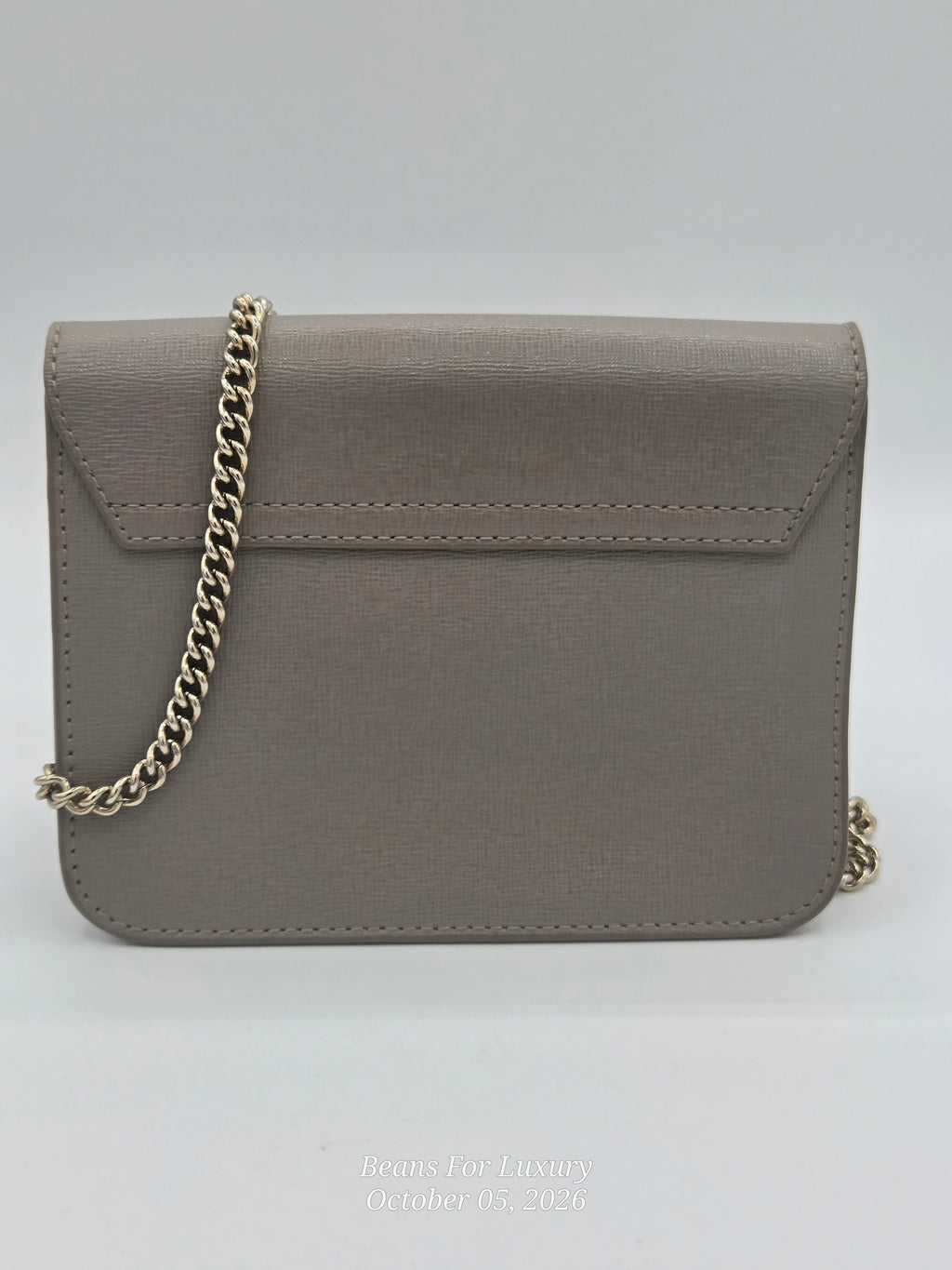 A taupe faux-saffiano leather crossbody bag with a gold chain strap, shown against a neutral light gray background.