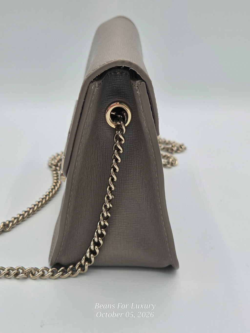 A taupe leather shoulder bag shown from a side angle, featuring a gold-tone chain strap and a gold grommet detail, set against a light gray background.