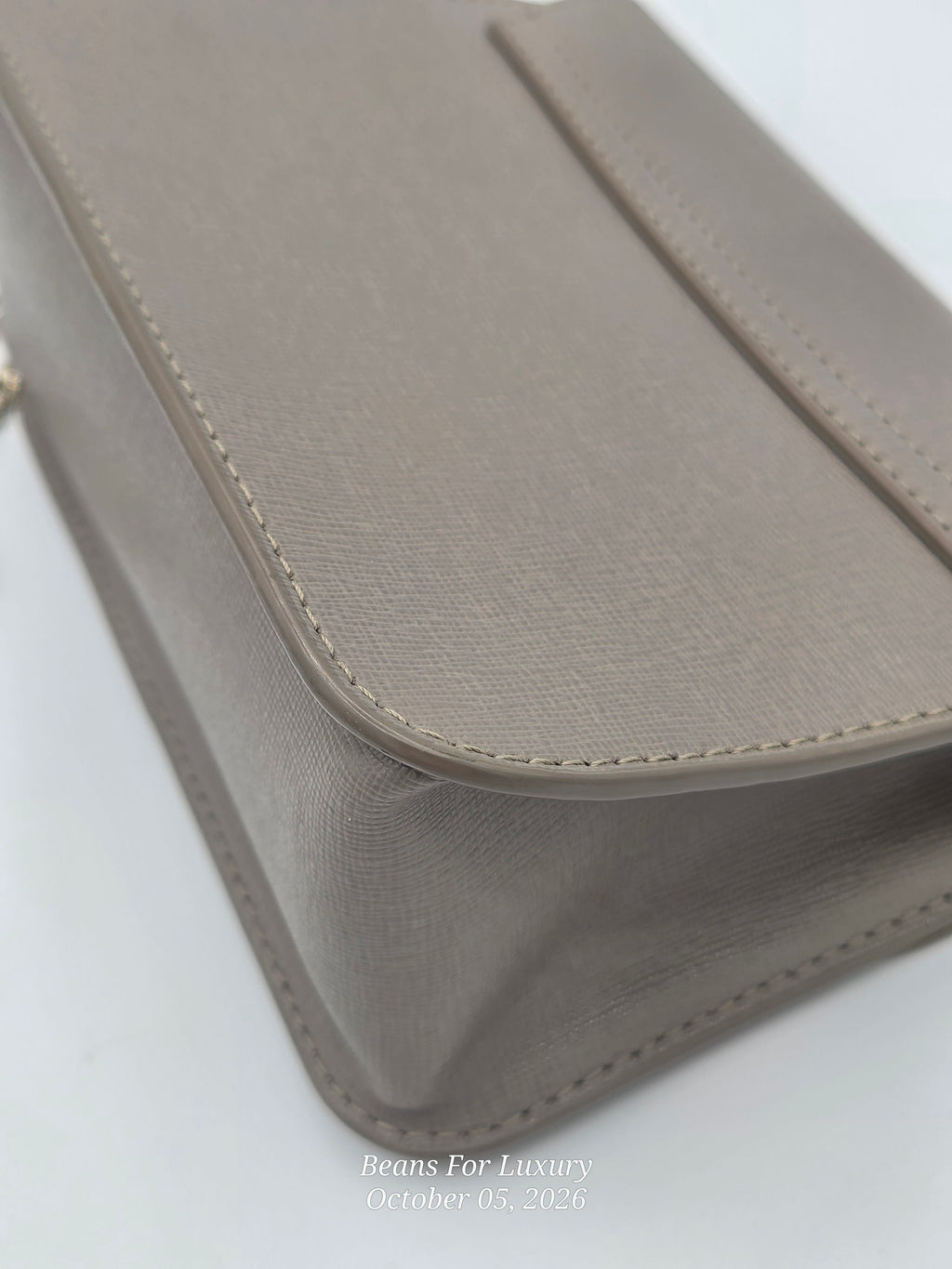 A close-up of a taupe leather handbag corner showing the textured grain, clean stitching, and a small gold ring clasp on a white background.