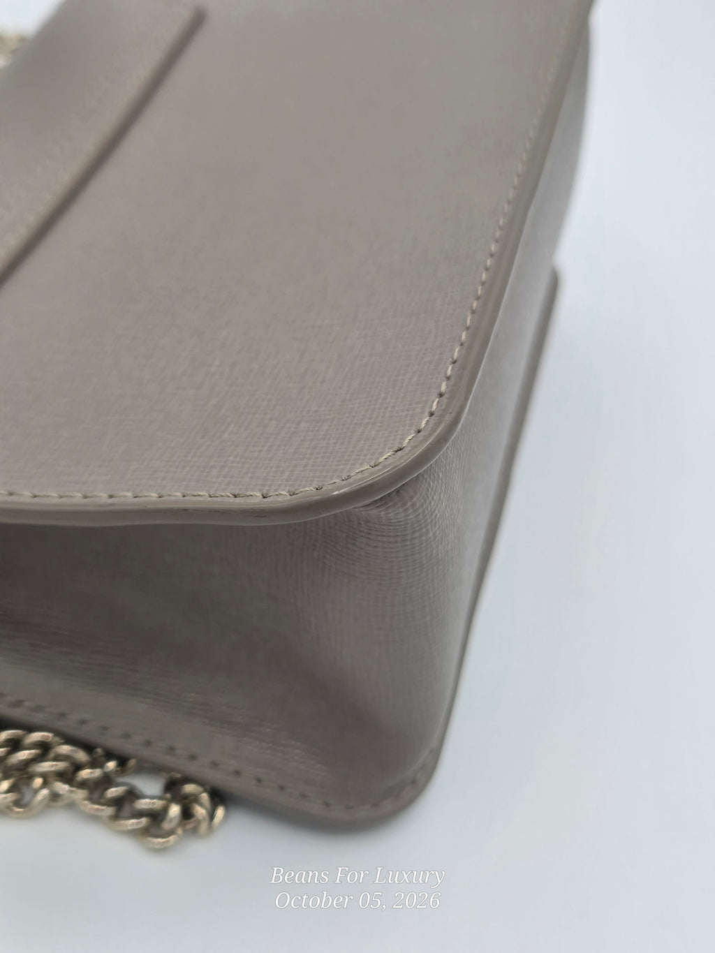 Close-up of a taupe leather shoulder bag with a gold chain strap on a white background.