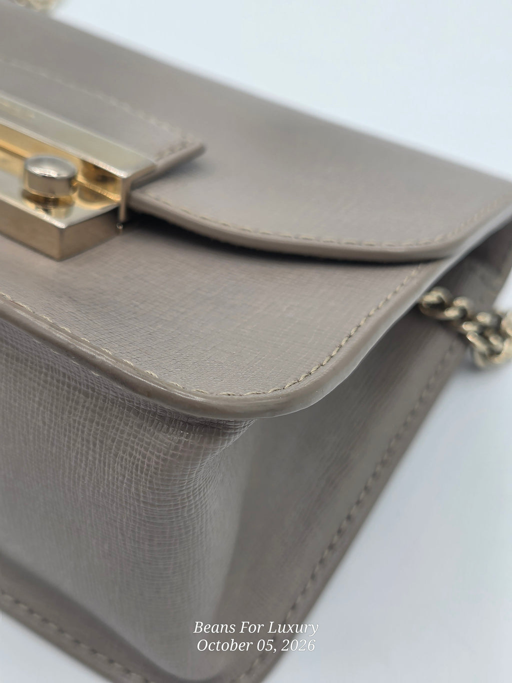 Close-up of a taupe textured leather handbag with gold-tone hardware and a chain strap, on a white background.