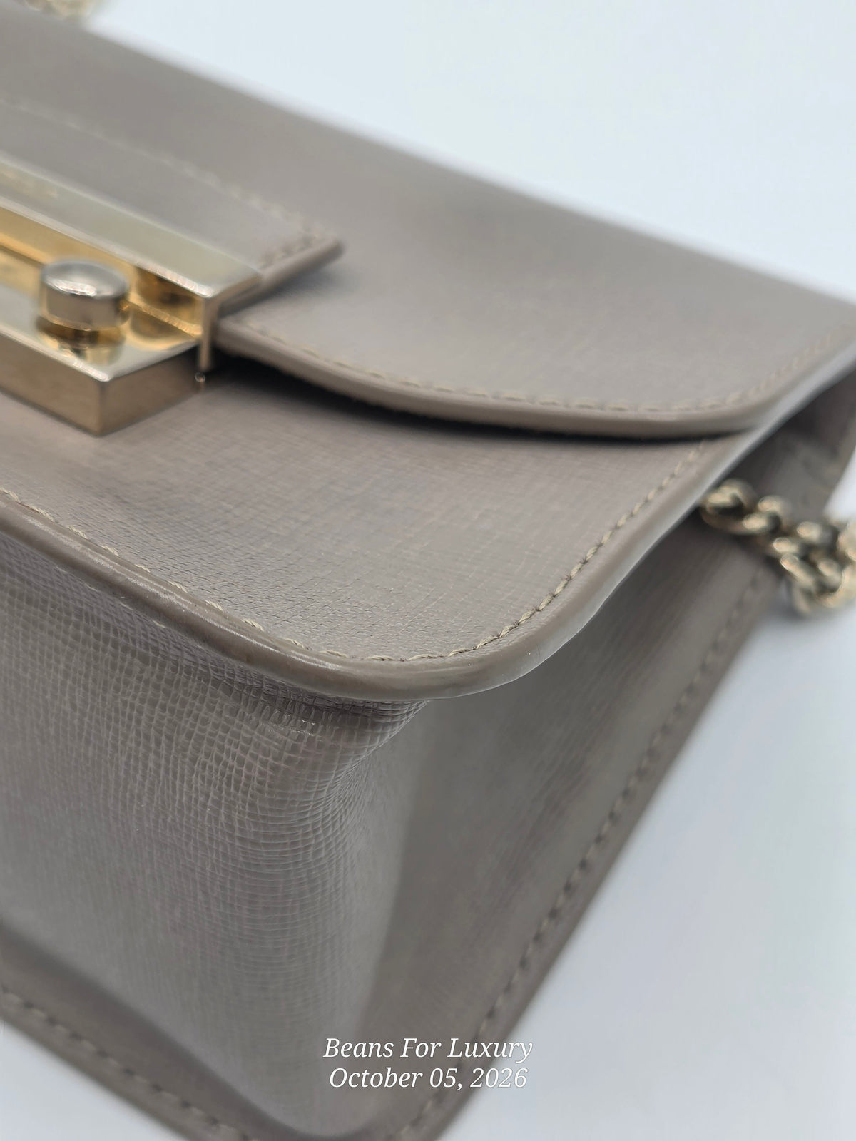 Close-up of a taupe textured leather handbag with gold-tone hardware and a chain strap, on a white background.