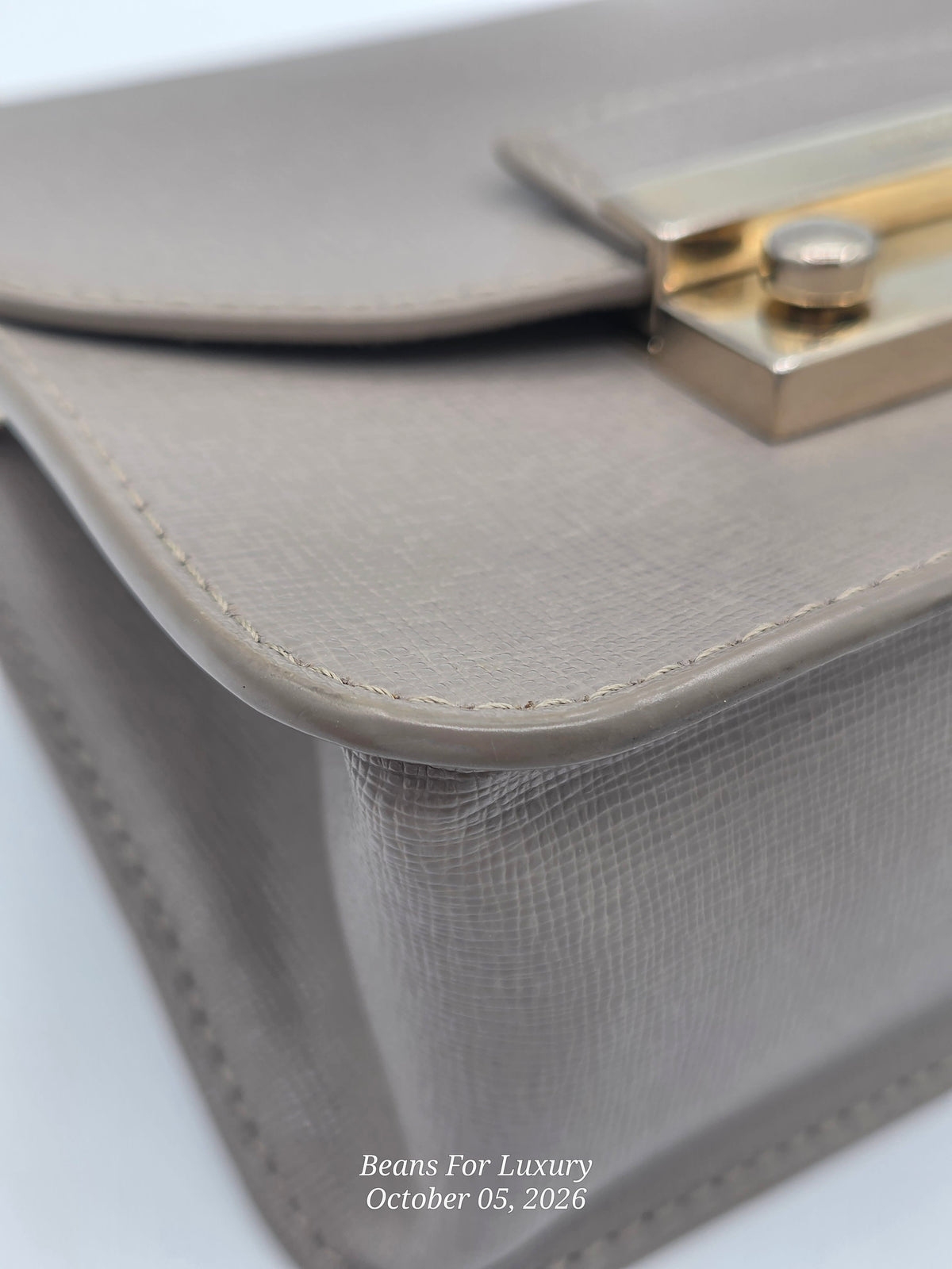 Close-up of a taupe leather handbag corner showing the textured leather grain, fine stitching, and a gold-tone metal clasp. A small watermark at the bottom reads Beans For Luxury October 05, 2026.
