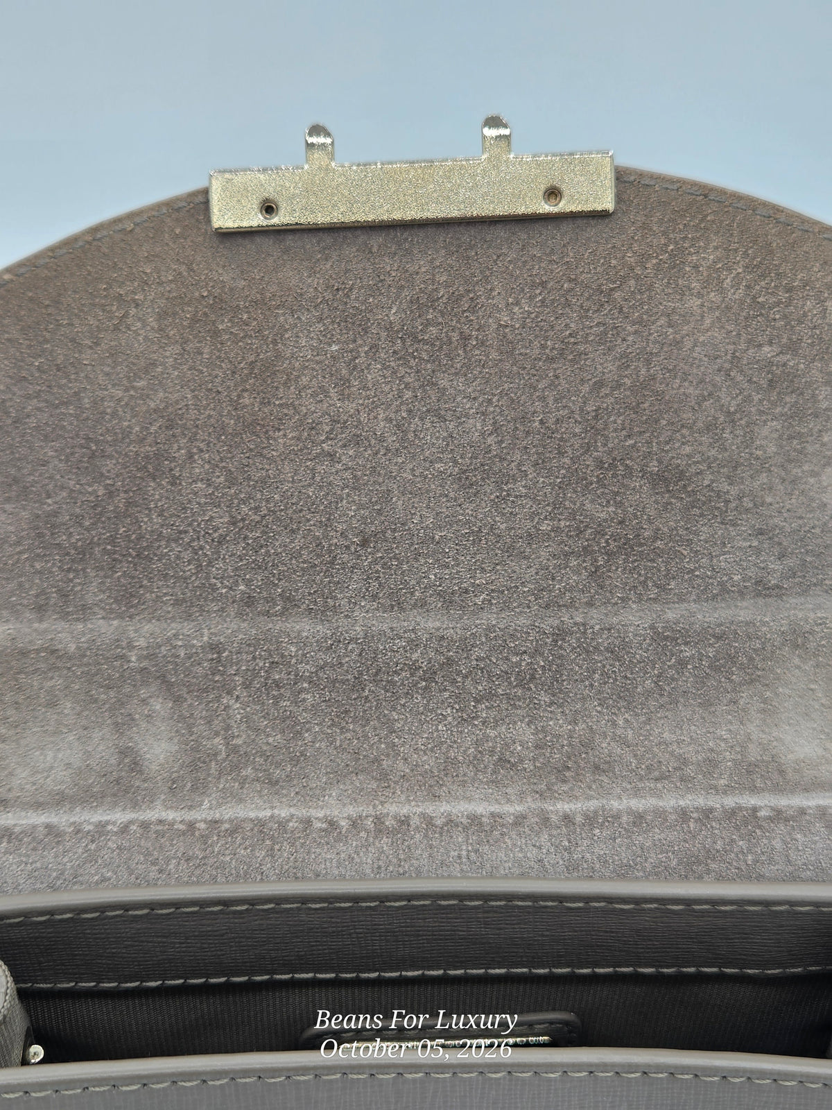 Close-up of the interior flap of a handbag in textured taupe-gray suede, featuring a gold-tone metal clip closure and dark gray lining.