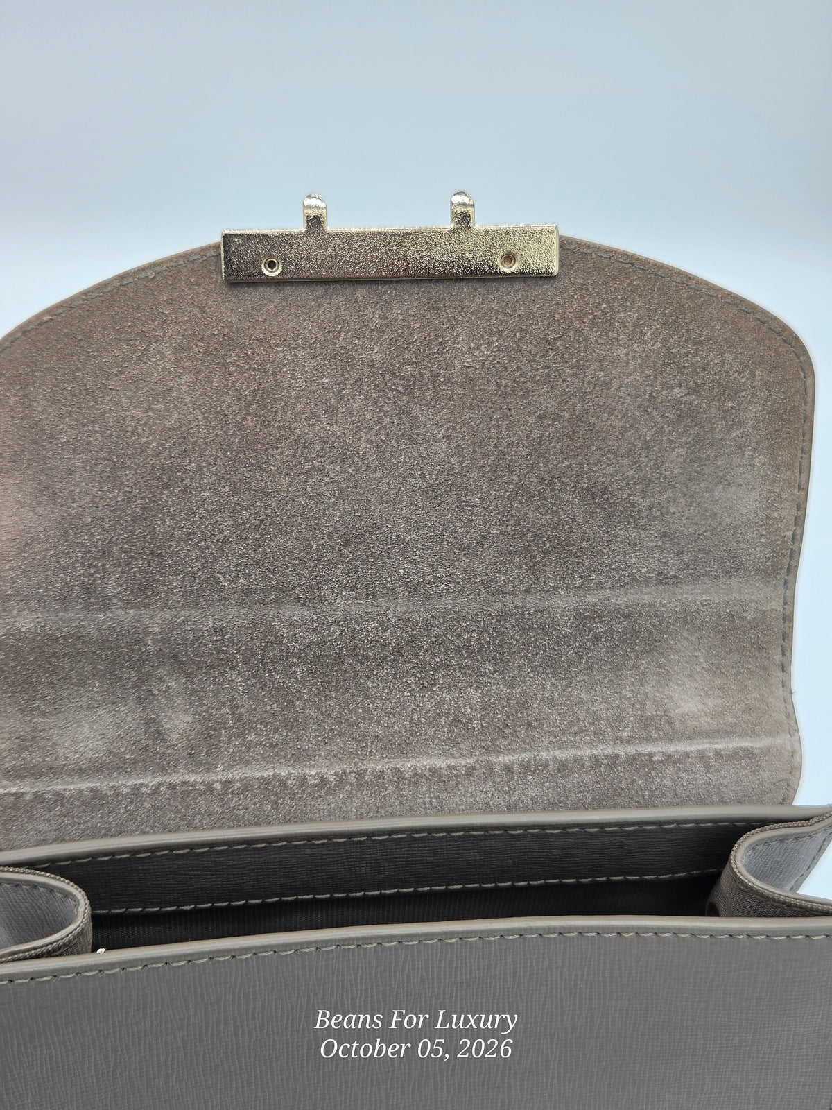 Close-up of the interior flap of a gray textured handbag, showing a silver-tone metal clasp and the inside lining, with a watermark reading 'Beans For Luxury' at the bottom.