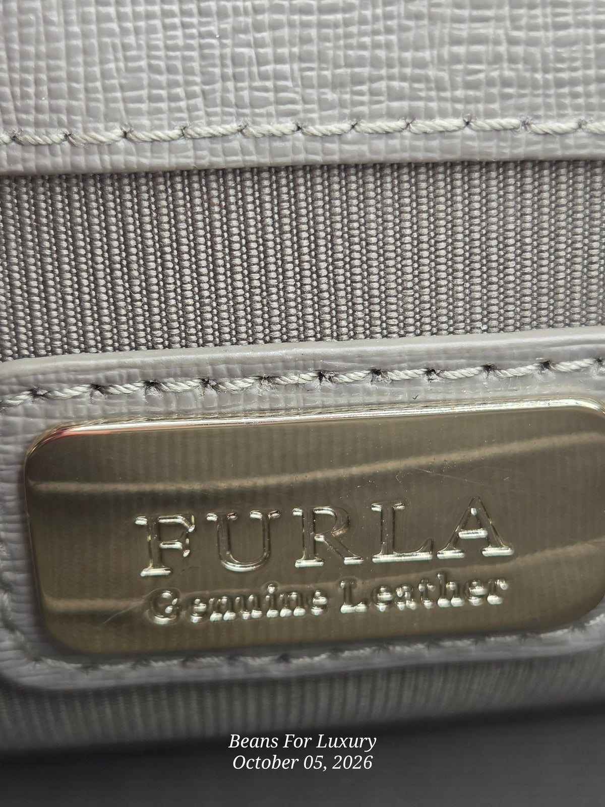 Close-up of a Furla leather product showing a brushed gold metal plaque with the embossed brand name 'FURLA Genuine Leather' on a textured light gray leather surface with a woven horizontal band.