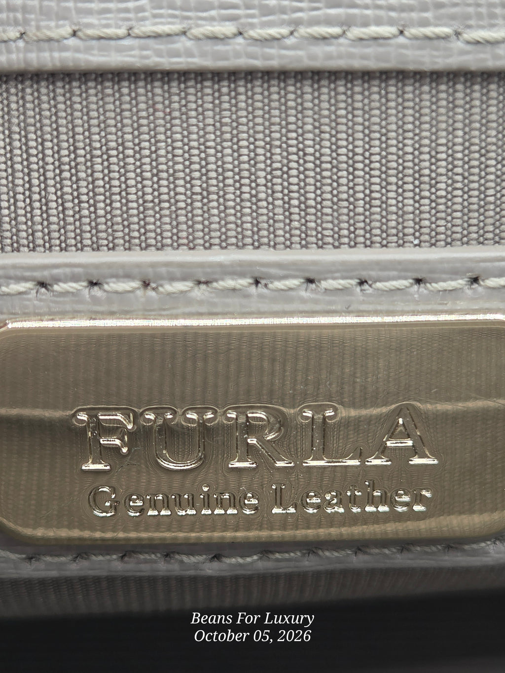 Close-up of a Furla leather product showing a brushed gold-tone metal plaque engraved with 'FURLA Genuine Leather' and light gray woven fabric with white stitching.