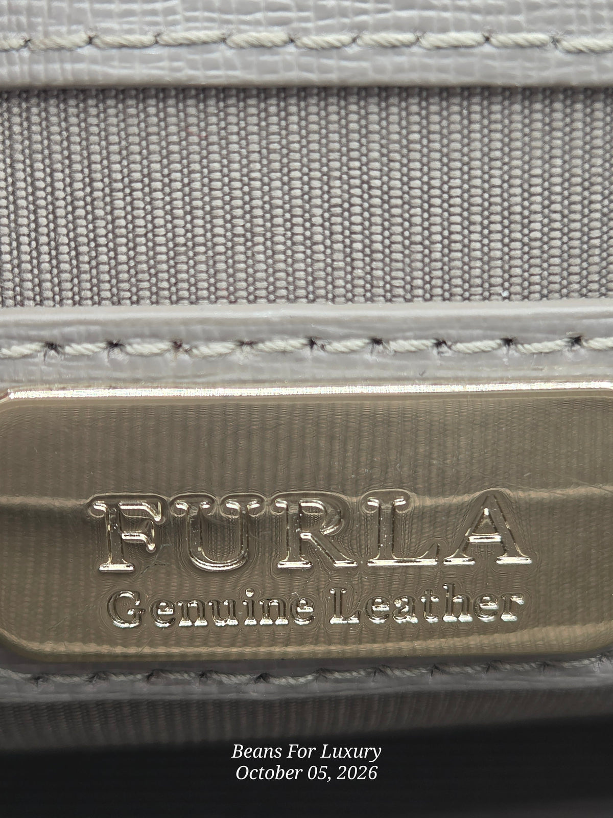 Close-up of a Furla leather product showing a brushed gold-tone metal plaque engraved with 'FURLA Genuine Leather' and light gray woven fabric with white stitching.