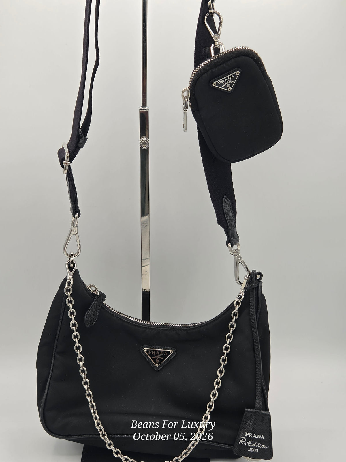 Black Prada shoulder bag with a matching small crossbody pouch, displayed on a stand against a light gray background. The bags feature silver-tone chain straps and the signature triangular Prada logo.