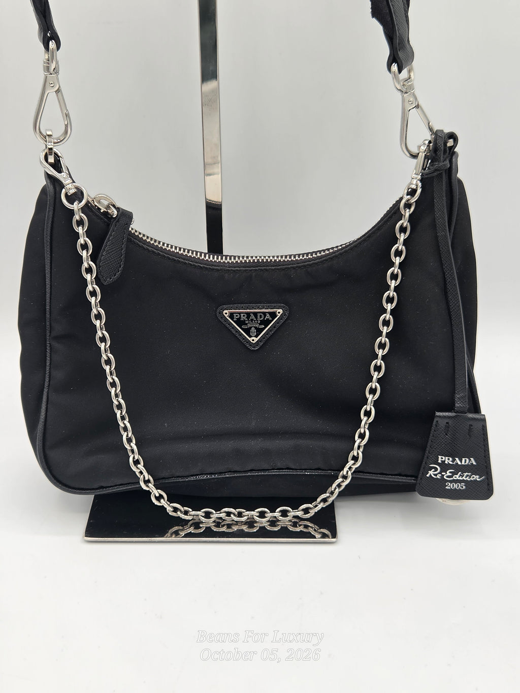 A black Prada shoulder bag made of nylon fabric, featuring a silver-tone chain strap draped across the front, a silver-tone metal triangle logo plaque, and a black leather hangtag on a display stand.