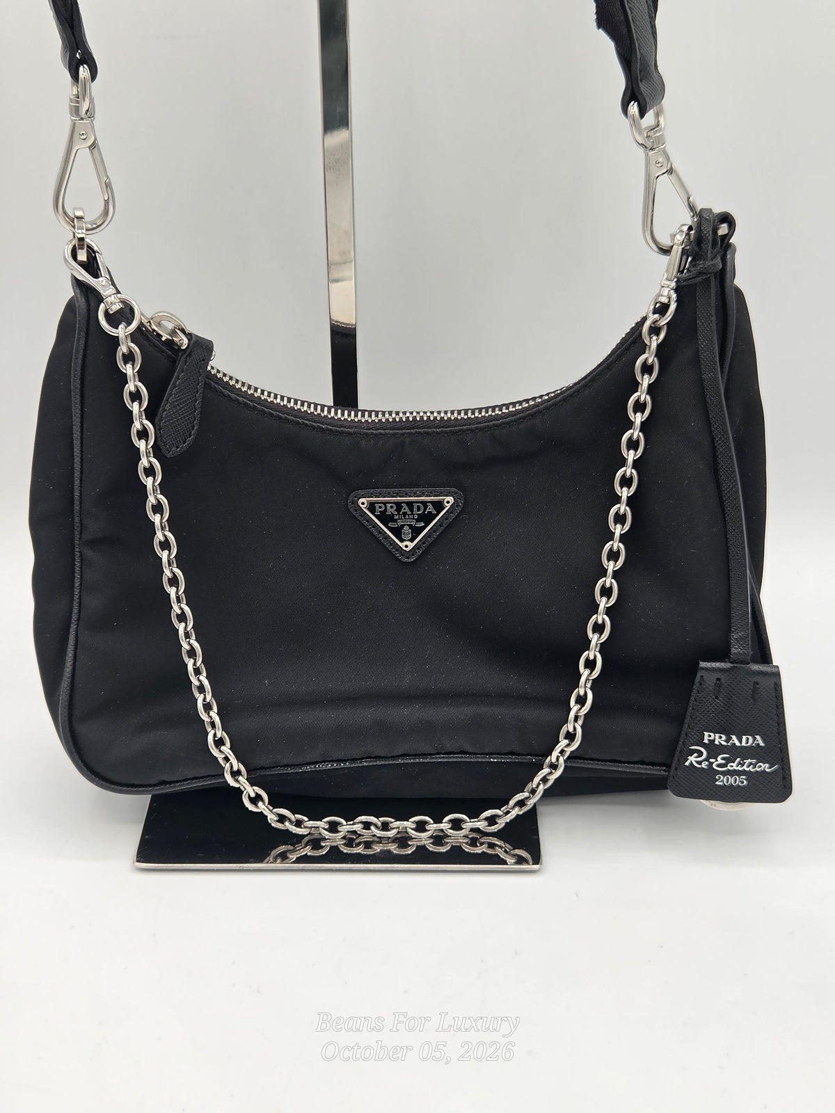 A black Prada shoulder bag made of nylon fabric, featuring a silver-tone chain strap draped across the front, a silver-tone metal triangle logo plaque, and a black leather hangtag on a display stand.