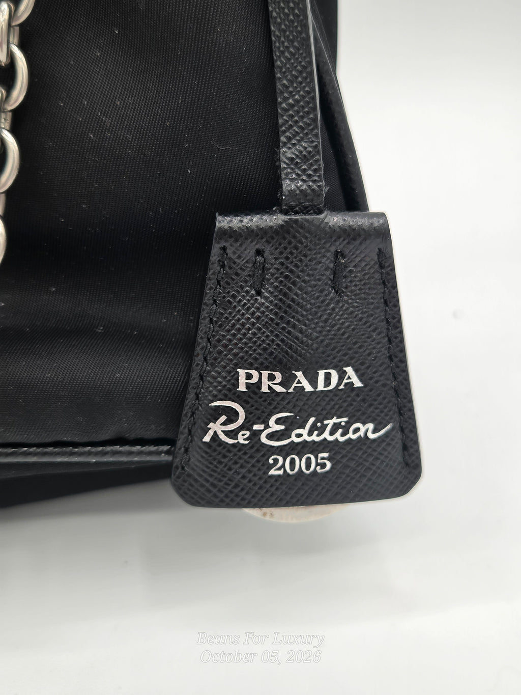 Close-up of a black Prada Re:Edition 2005 handbag tag with silver-tone lettering on a textured leather tab, attached to a black fabric bag body with a chain strap visible on the left.