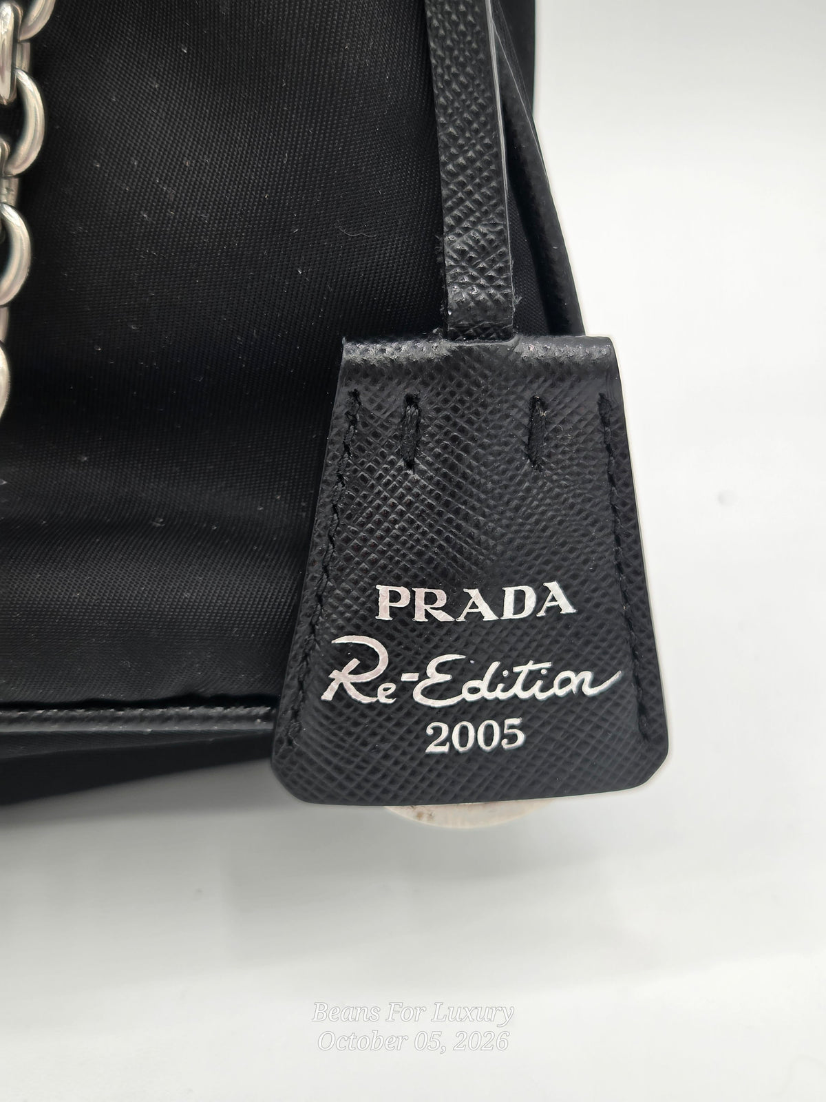 Close-up of a black Prada Re:Edition 2005 handbag tag with silver-tone lettering on a textured leather tab, attached to a black fabric bag body with a chain strap visible on the left.