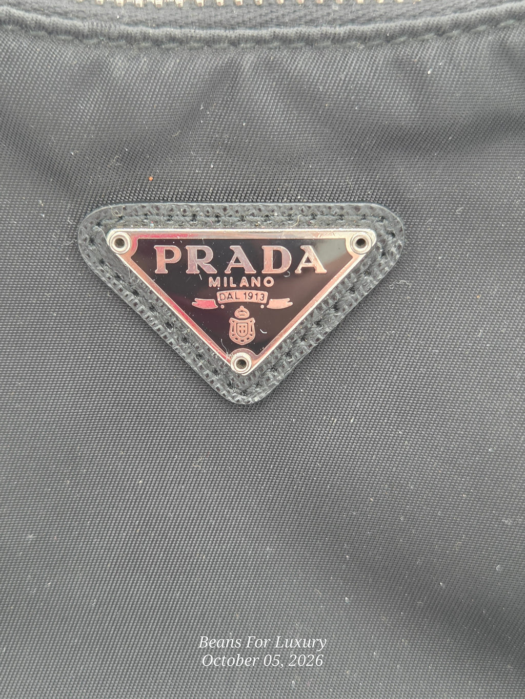 Close-up of a Prada Milano triangle metal logo plaque on a dark gray textured fabric surface, with a small 'Beats For Luxury' watermark at the bottom.