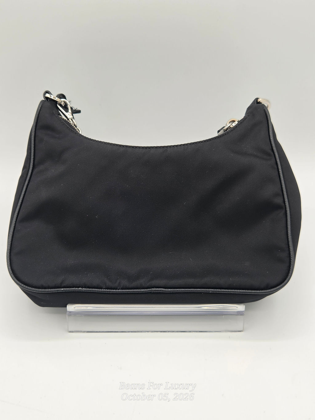 A black nylon shoulder bag with silver-tone hardware, displayed on a clear acrylic stand against a light gray background.