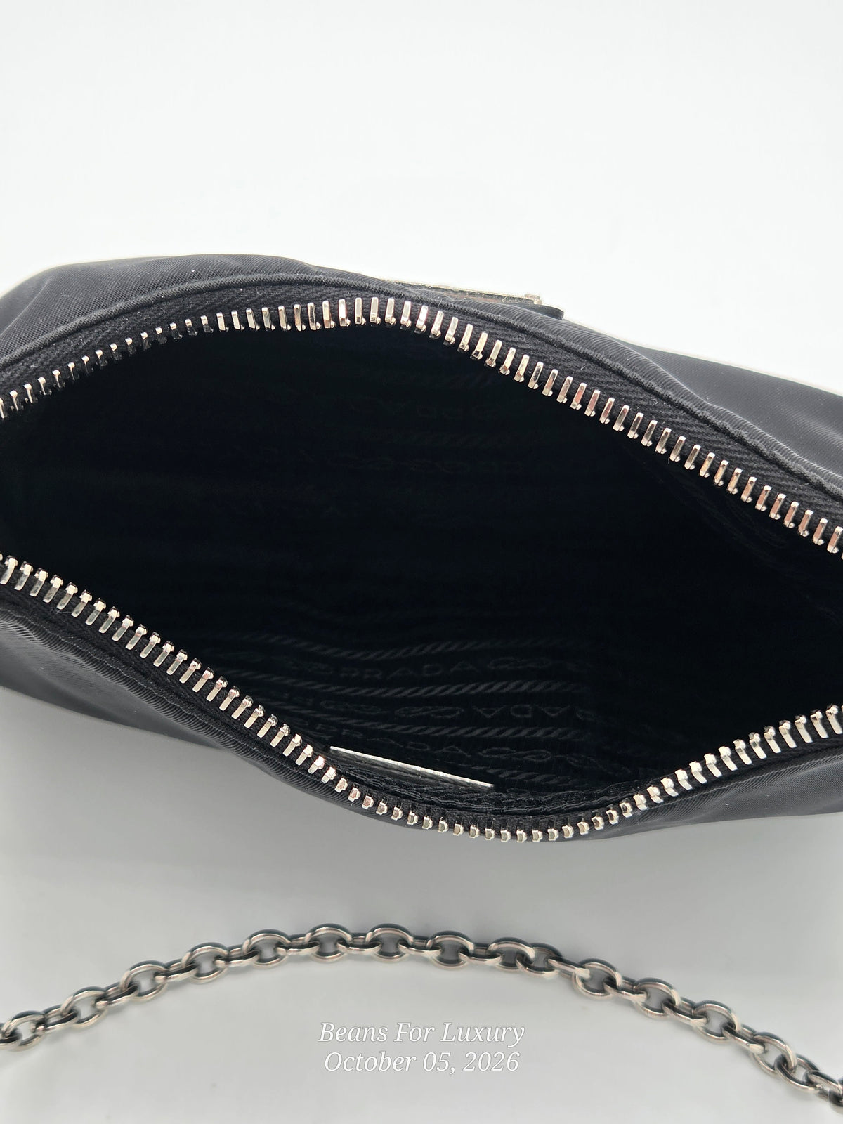 Close-up of an open black leather clutch or handbag showing the black interior lining and silver-tone zipper. A silver chain strap is visible at the bottom against a light gray background.