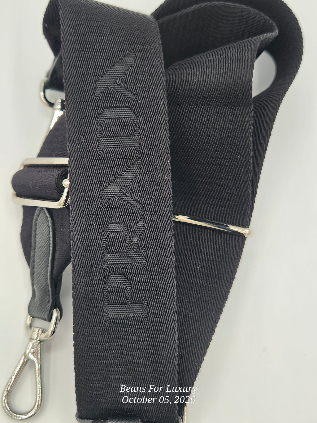 A close-up of a black fabric shoulder strap with silver-tone metal hardware, including a buckle, adjustment loop, and snap hook. The strap features a debossed vertical brand name that appears to read 'BEYOND'.