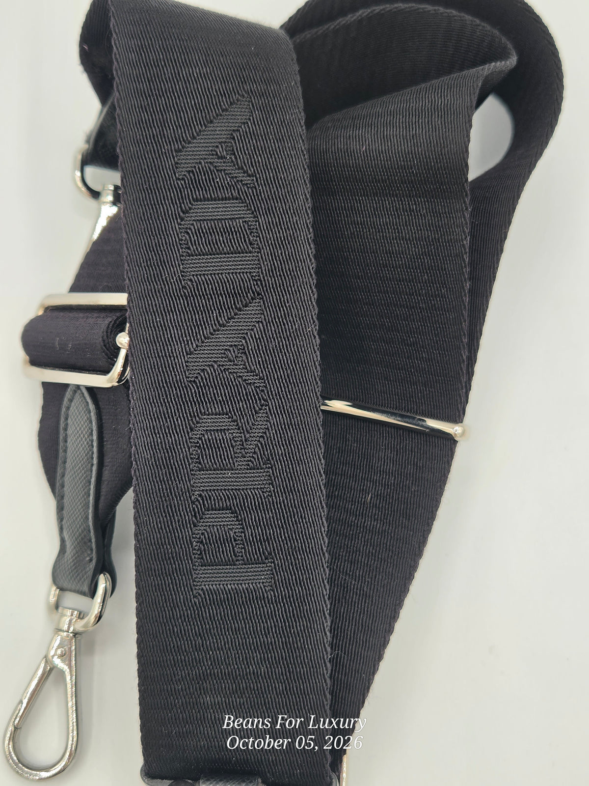 A close-up of a black fabric shoulder strap with silver-tone metal hardware, including a buckle, adjustment loop, and snap hook. The strap features a debossed vertical brand name that appears to read 'BEYOND'.