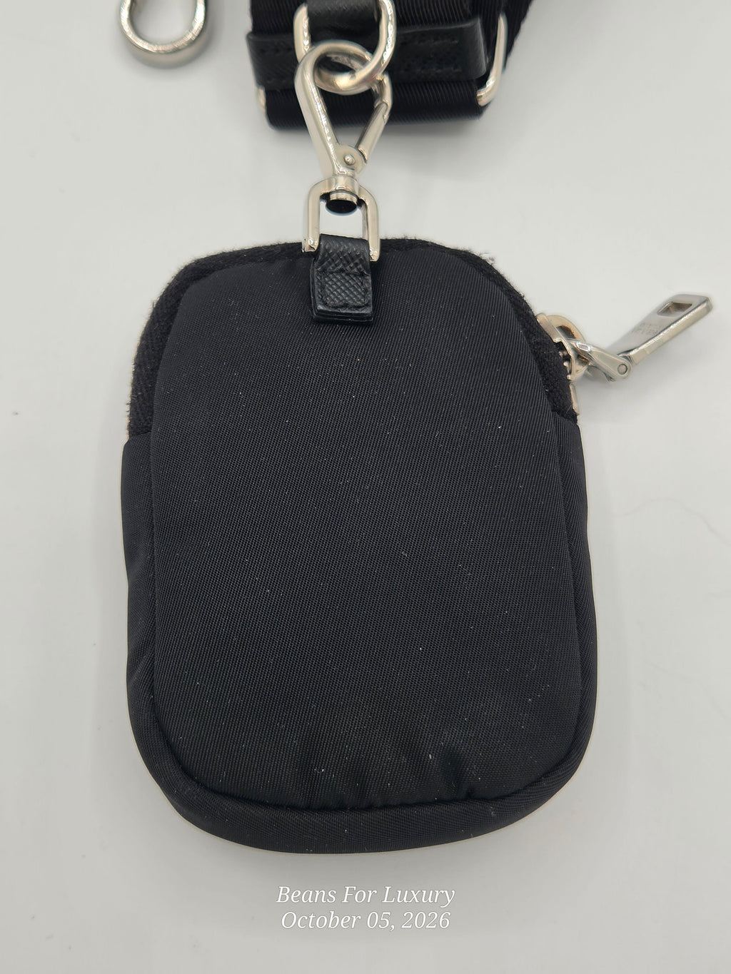 A small black fabric wristlet or pouch with a silver metal clasp and strap, photographed on a light gray background. A small watermark reading 'Beans For Luxury October 05, 2026' appears near the bottom.