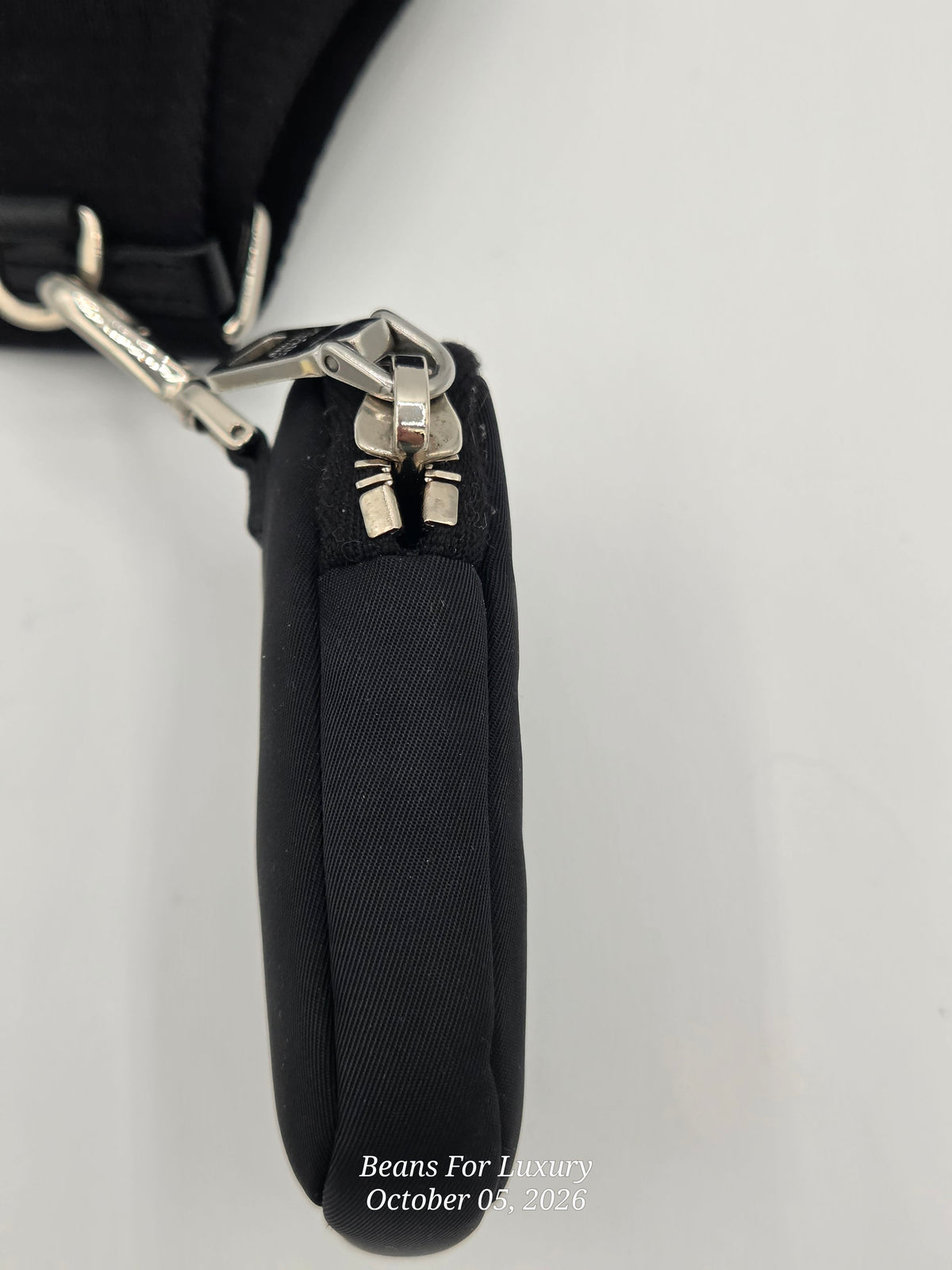 Close-up of a black fabric wristlet or pouch with a silver metal zipper and hardware, shown on a plain light background. A small watermark at the bottom reads Beans For Luxury October 05, 2026.