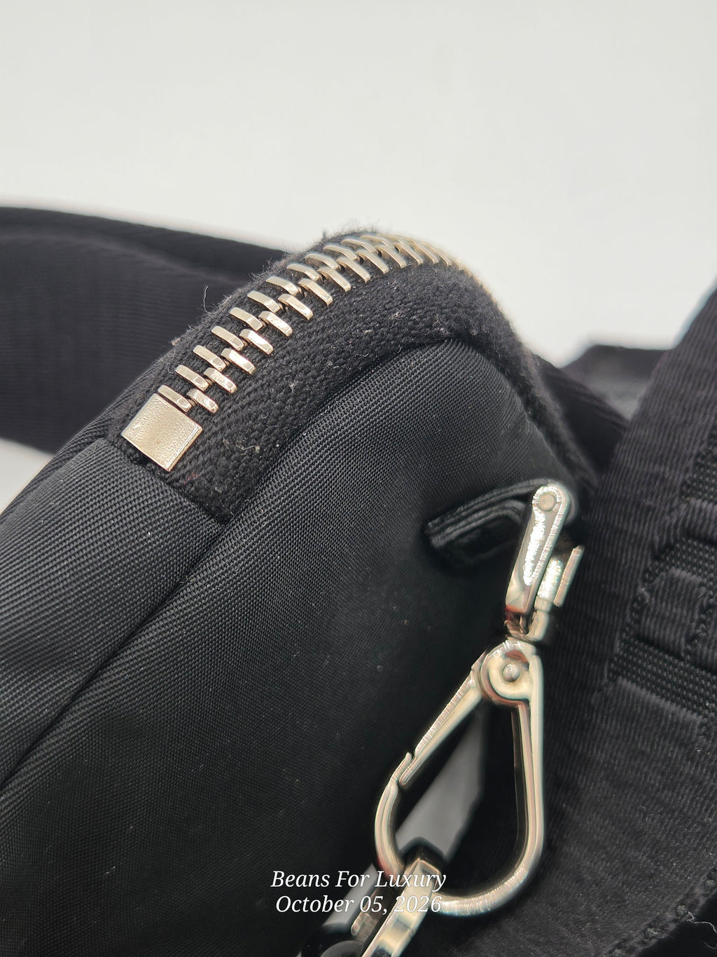 Close-up of a black fabric bag or pouch showing a silver metal zipper and a silver carabiner clip attachment, with a small Beans For Luxury watermark at the bottom.