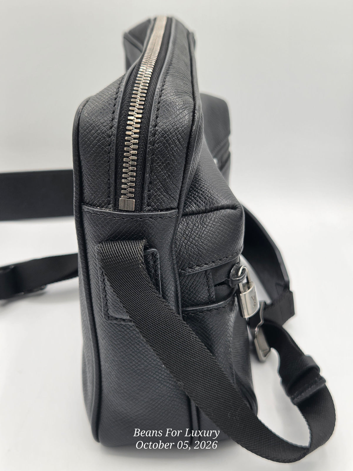 A black textured leather crossbody bag with a silver zipper and adjustable shoulder strap, shown from a side profile view on a light gray background.