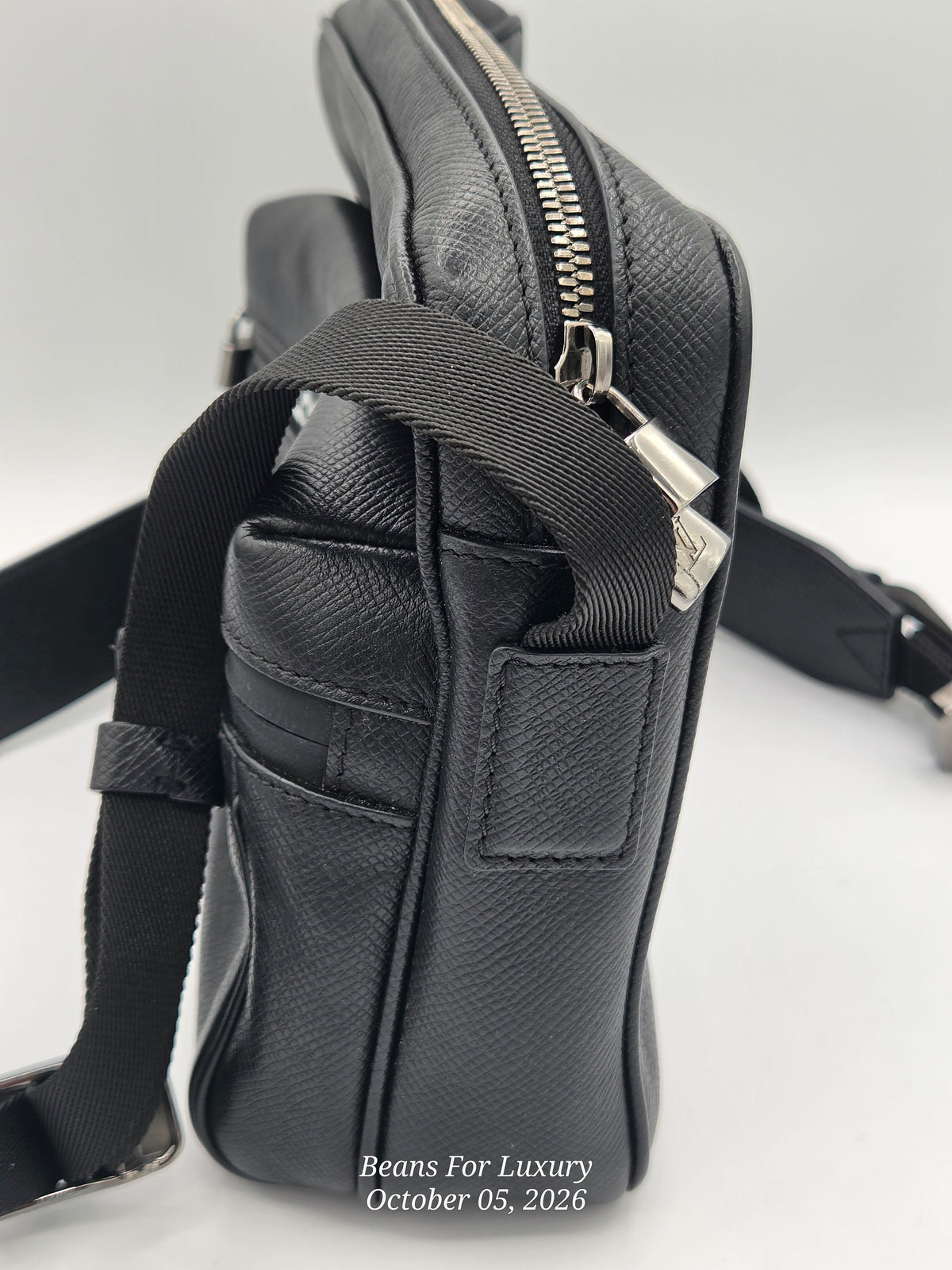 A close-up side view of a black textured leather crossbody bag with a black webbing strap and silver zipper hardware, photographed on a light gray background.