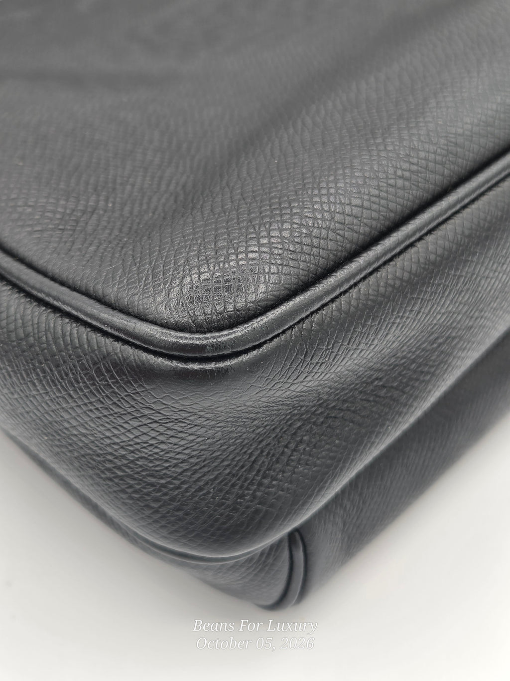 Close-up of a black textured leather handbag corner showing the pebbled leather grain and stitched edge on a white background.