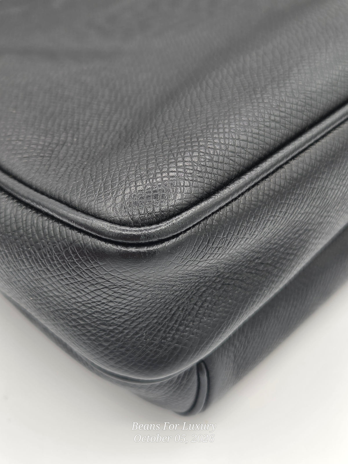 Close-up of a black textured leather handbag corner showing the pebbled leather grain and stitched edge on a white background.