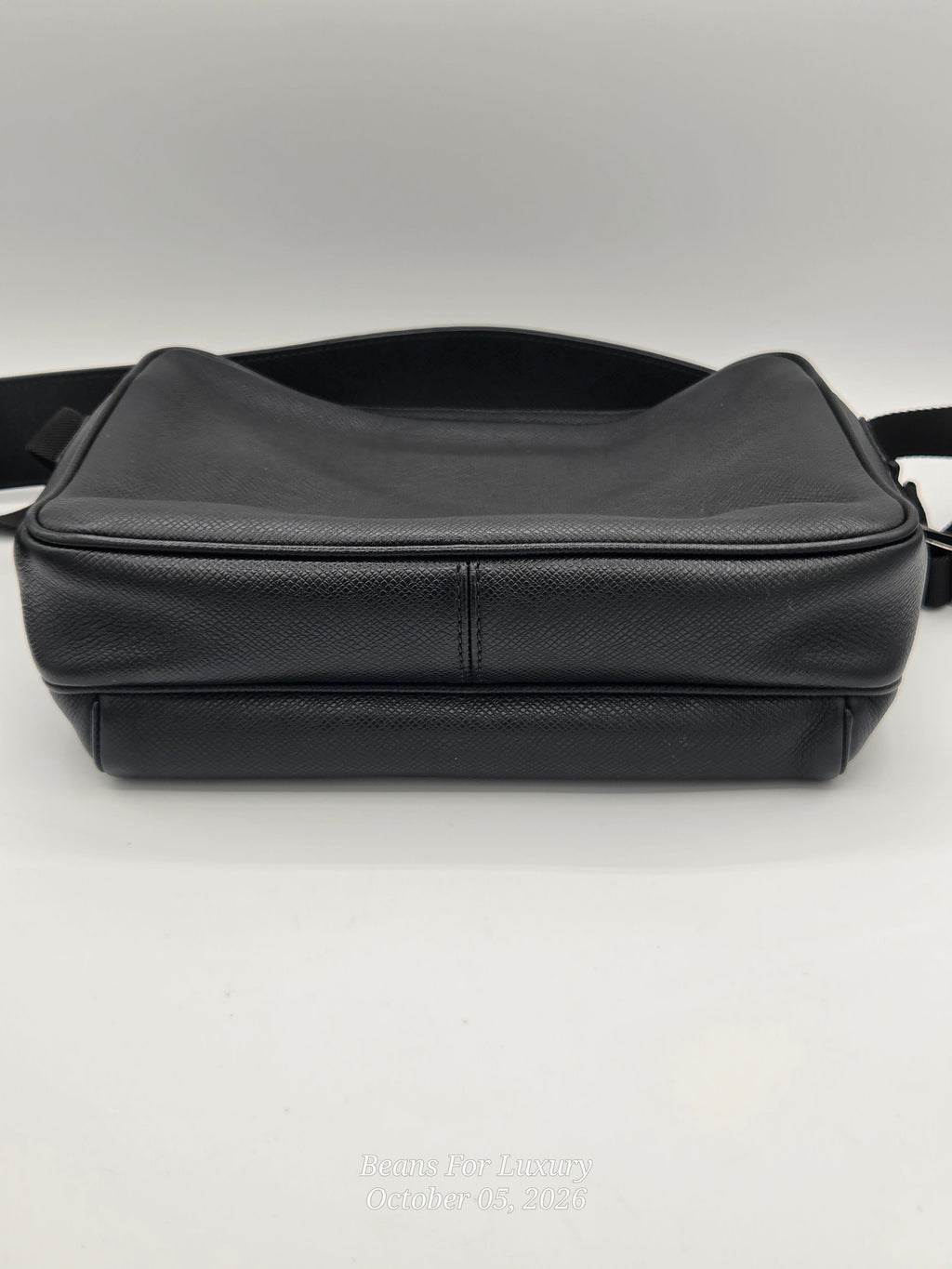 A black textured leather messenger bag with a wide shoulder strap, photographed from the side against a neutral light gray background.