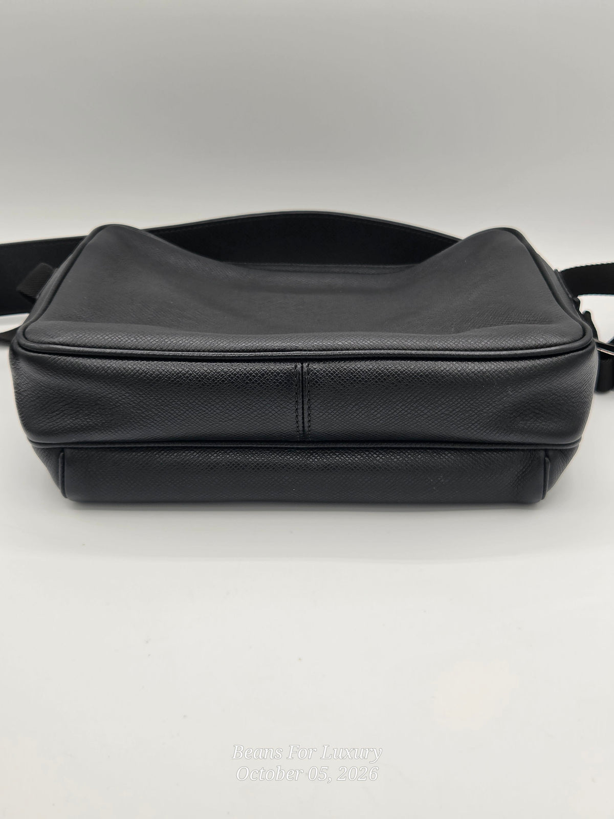 A black textured leather messenger bag with a wide shoulder strap, photographed from the side against a neutral light gray background.