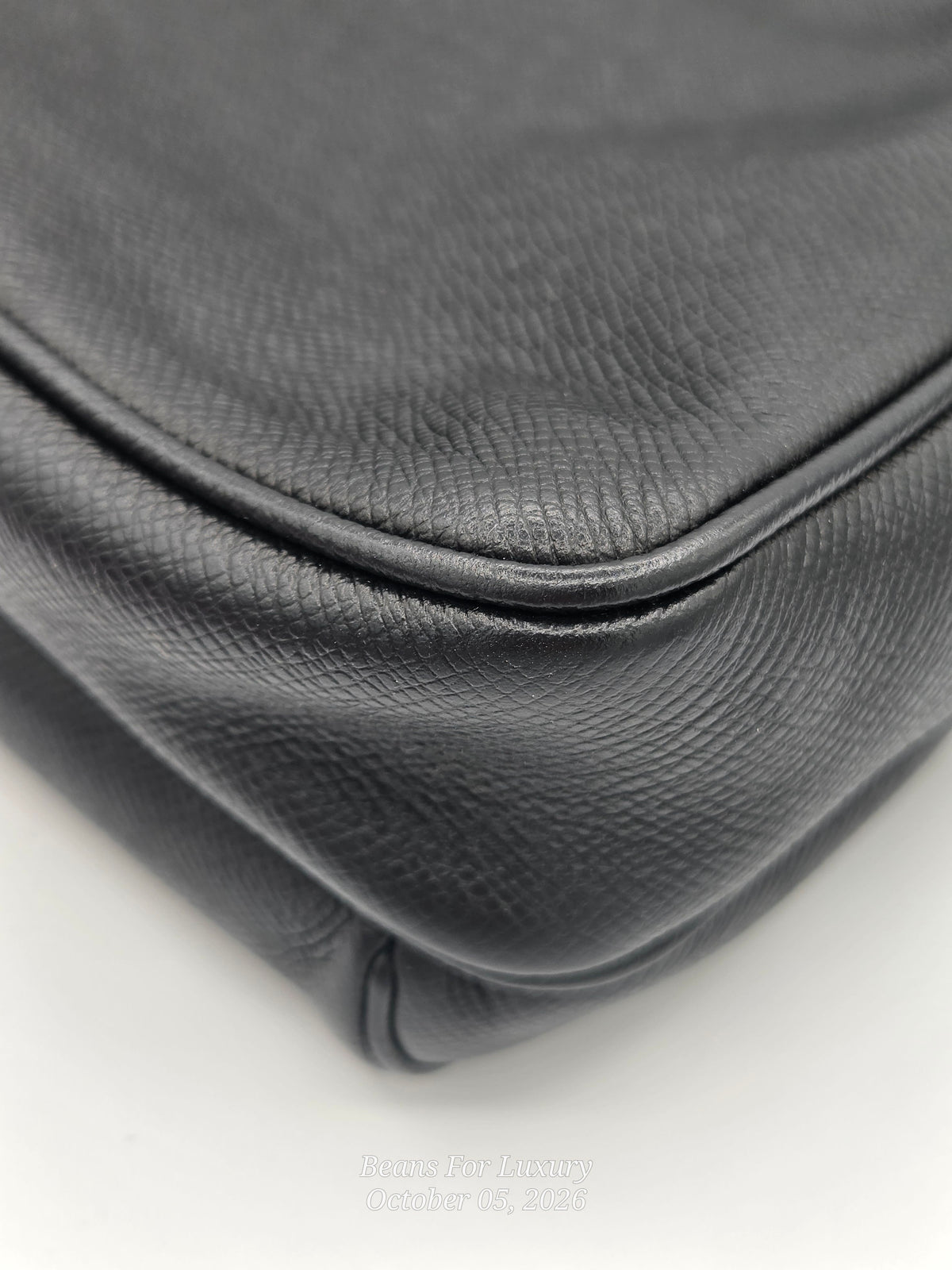 Close-up detail shot of a black pebbled leather handbag corner, showing the texture and stitching on a white background.